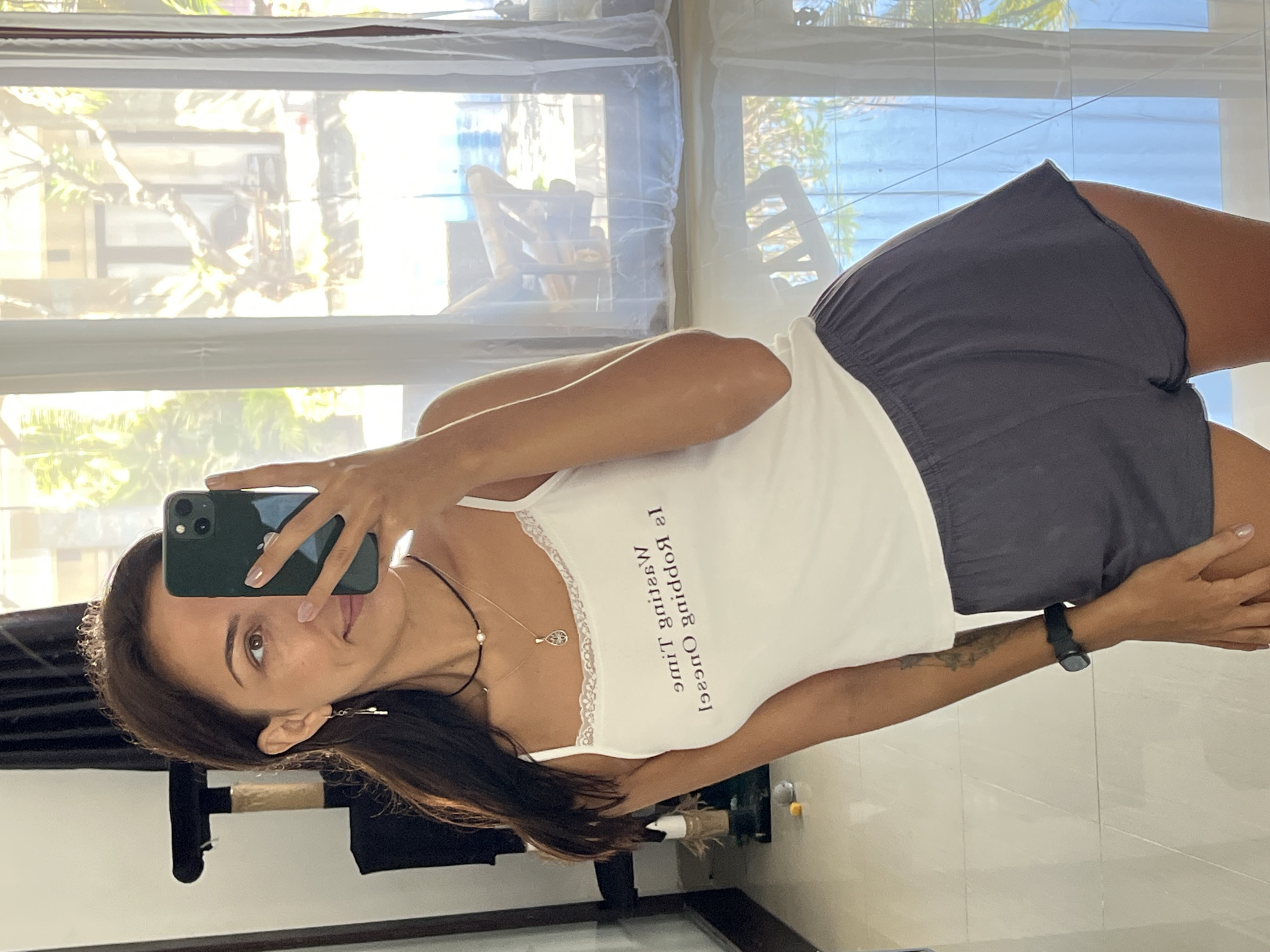 A woman is taking a selfie indoors in Unknown, Indonesia. She is wearing a white tank top with black shorts. She is holding a smartphone with her left hand. The background is a window showing trees and buildings.