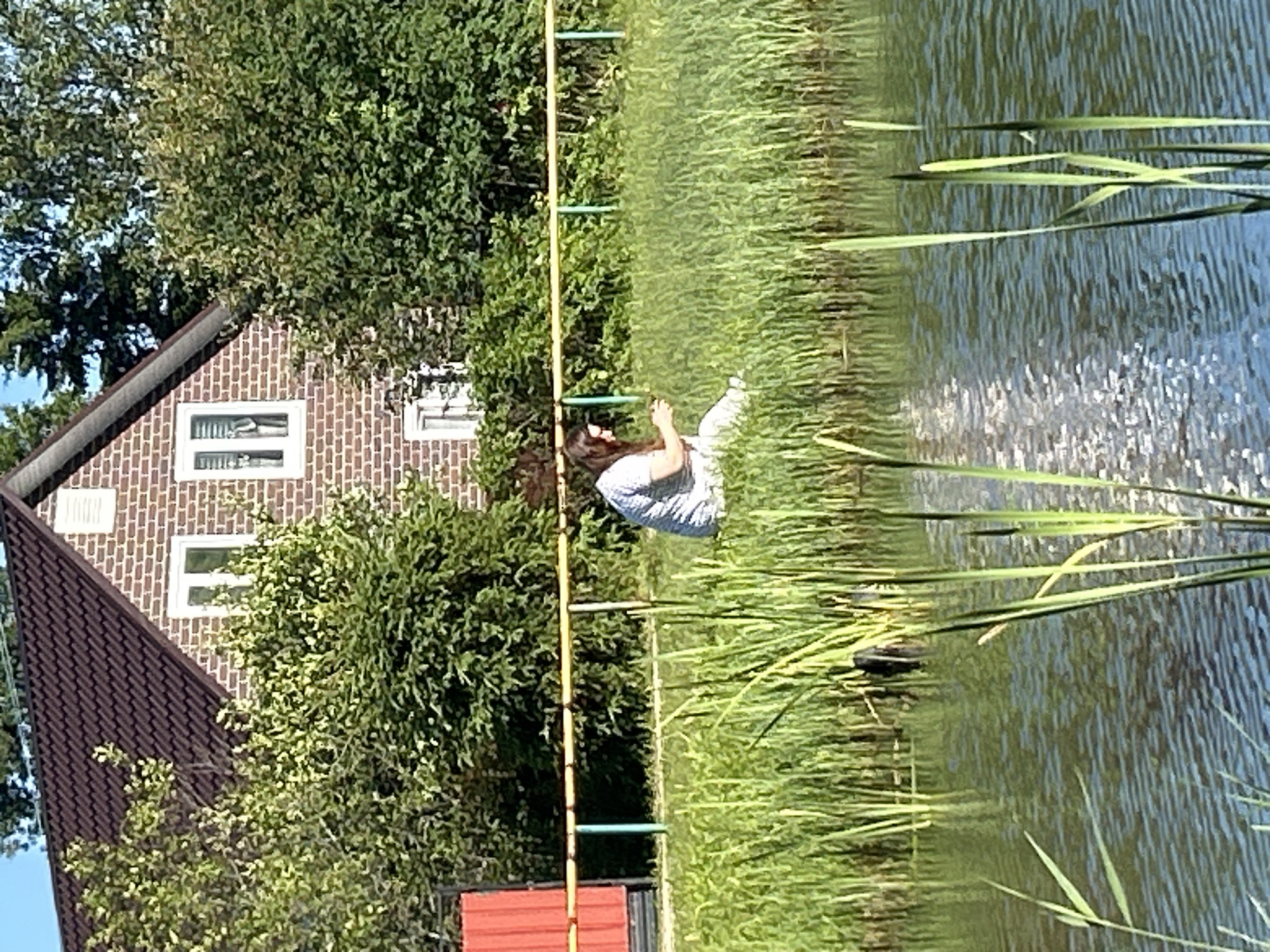 A woman is sitting in the tall green grass on the bank of a body of water. She is wearing a light-colored, patterned shirt and pants and appears to be looking at her phone. Behind her, there is a yellow railing with green posts, and further back is a brick house with a dark roof and trees. The water in front of her is rippled, reflecting the sunlight. The scene appears to be in a suburban or rural area.