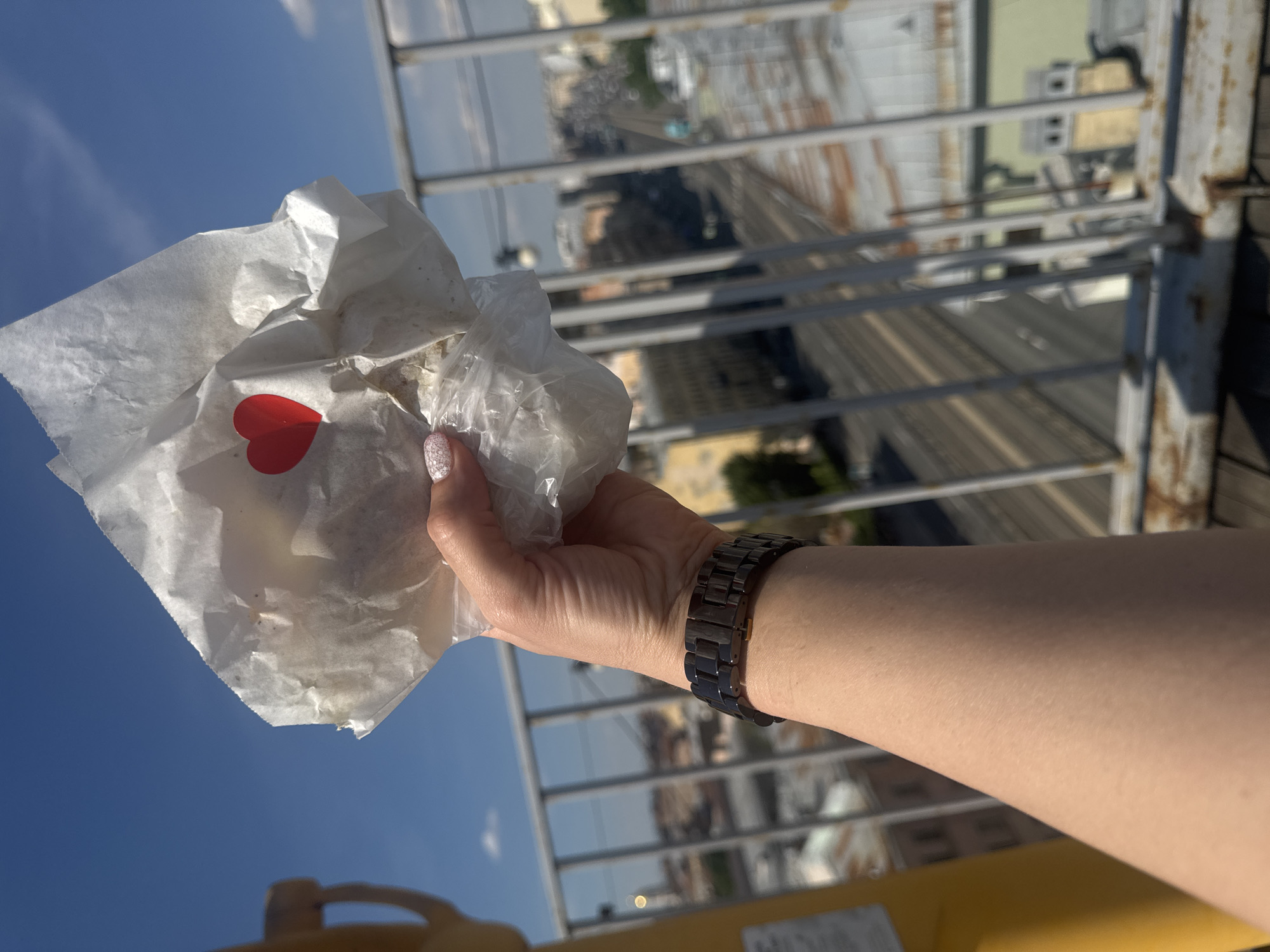 A hand holding a wrapped food item, adorned with a small red heart sticker, is visible against a bright blue sky and a cityscape. The food is wrapped in white paper and plastic. The hand has light skin and is wearing a dark metal watch on the wrist. In the background, a metal railing separates the viewer from a view of rooftops, streets, and buildings in Saint Petersburg, Russia. The overall lighting suggests a sunny day.