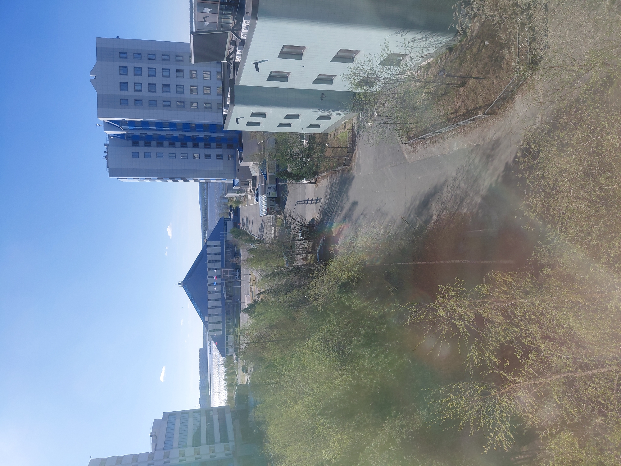 The image presents an elevated view of an urban landscape in Nadym, Russia, on a bright, sunny day during springtime. The sky is a clear, vibrant blue with a few wispy clouds. Strong sunlight creates distinct shadows and prominent lens flares across the frame, indicating a clear, daytime scene, likely late morning or early afternoon.
The scene is dominated by several buildings and abundant greenery. On the left, two tall, light-gray high-rise buildings feature multiple rectangular windows and striking vertical blue accents, possibly reflective glass or painted panels. To their right, a smaller, light teal-colored building with uniquely shaped, slightly recessed windows and visible air conditioning units on its roof stands. In the distant background, a large building with a dark, steeply sloped roof is visible, along with other urban structures.
An asphalt road or path meanders through the midground, bordered by numerous trees beginning to leaf out with fresh, light green foliage, characteristic of early spring. Beyond the buildings and trees, a wide body of water, likely a river or lake, glistens under the sun, reflecting the sky. Some metal railings are visible near the water's edge.
No people or specific activities are discernible in the static scene. There is no visible text that can be clearly read. The overall impression is a modernizing urban environment amidst a natural setting, bathed in sunlight.