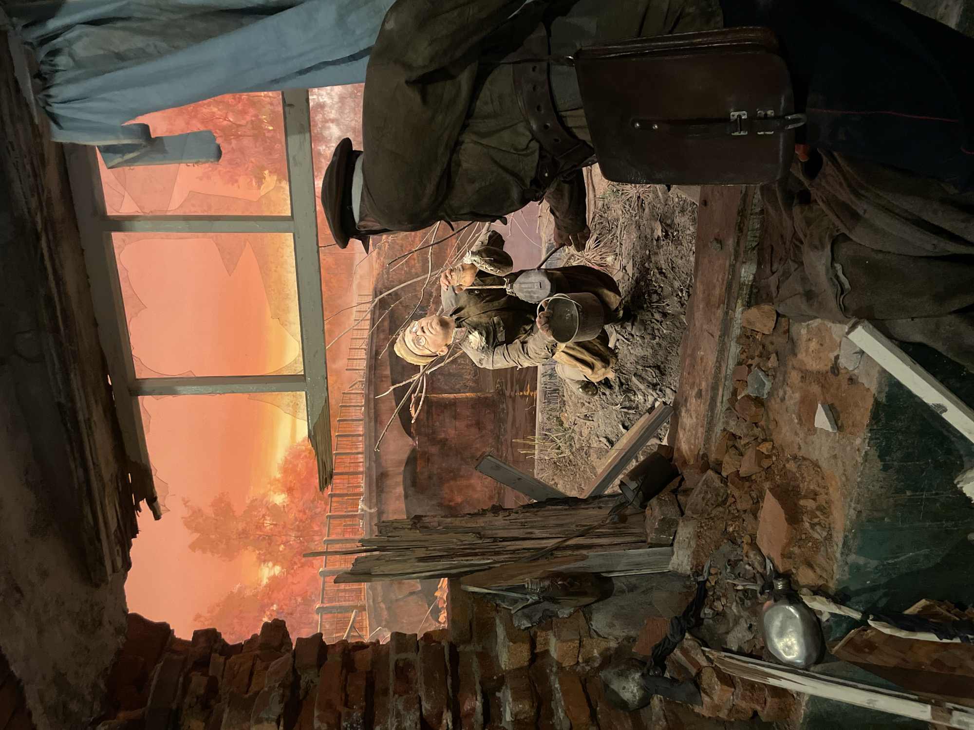 The image depicts a diorama, possibly located in the city of Moscow, Russia, recreating a scene of wartime devastation. A building is heavily damaged, with broken brickwork and shattered windows revealing a fiery, possibly sunset-lit, sky. Two uniformed figures, likely mannequins representing soldiers, are central. One is partially visible from the back, wearing a dark uniform with a brown leather satchel. The other soldier is depicted in a crouched position, holding containers. Debris, including wooden beams and what appears to be personal items like canteens, are scattered throughout the scene, emphasizing the destruction. The diorama attempts to capture the atmosphere of conflict and destruction.