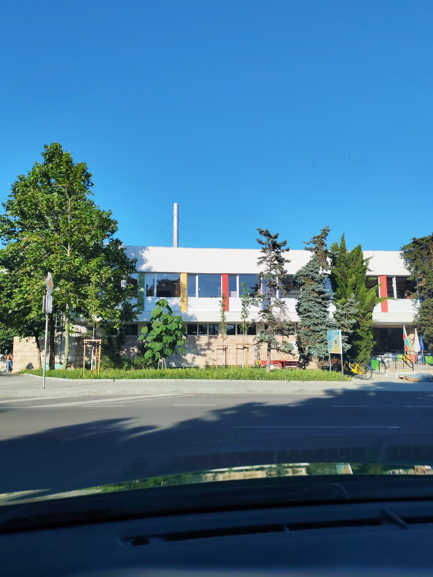 In the city of Burgas, Bulgaria, a modern, two-story building with large windows and colorful vertical accents stands beneath a clear blue sky. The building is bordered by a well-maintained green lawn and a sidewalk, with trees of varying sizes providing shade. A street with a marked crosswalk is visible in the foreground, with shadows from the trees cast across it. To the left of the building, a lone person walks on the sidewalk. On the right side of the building, a yellow bicycle is parked near a sign and two Bulgarian flags are displayed. A chimney or vent pipe extends from the roof of the building. The overall scene depicts a bright, sunny day in a well-lit urban environment.