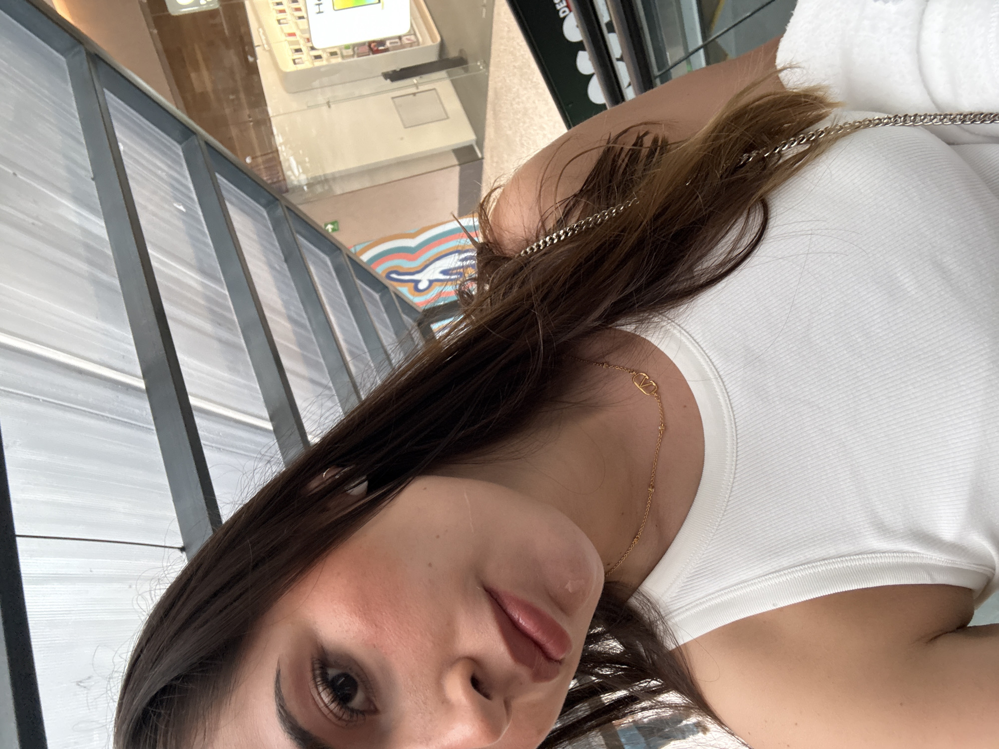 A young woman is facing the camera wearing a white tank top and a gold necklace. She has long brown hair. She appears to be standing on a balcony or elevated walkway with a metal and glass barrier. Below her, visible through the barrier, is a retail space with display cases. The photograph's location is inferred from the city of San Bernardino Tlaxcalancingo, Mexico. Specific environmental details, actions, interactions, and subject information are not discernible from the image provided.