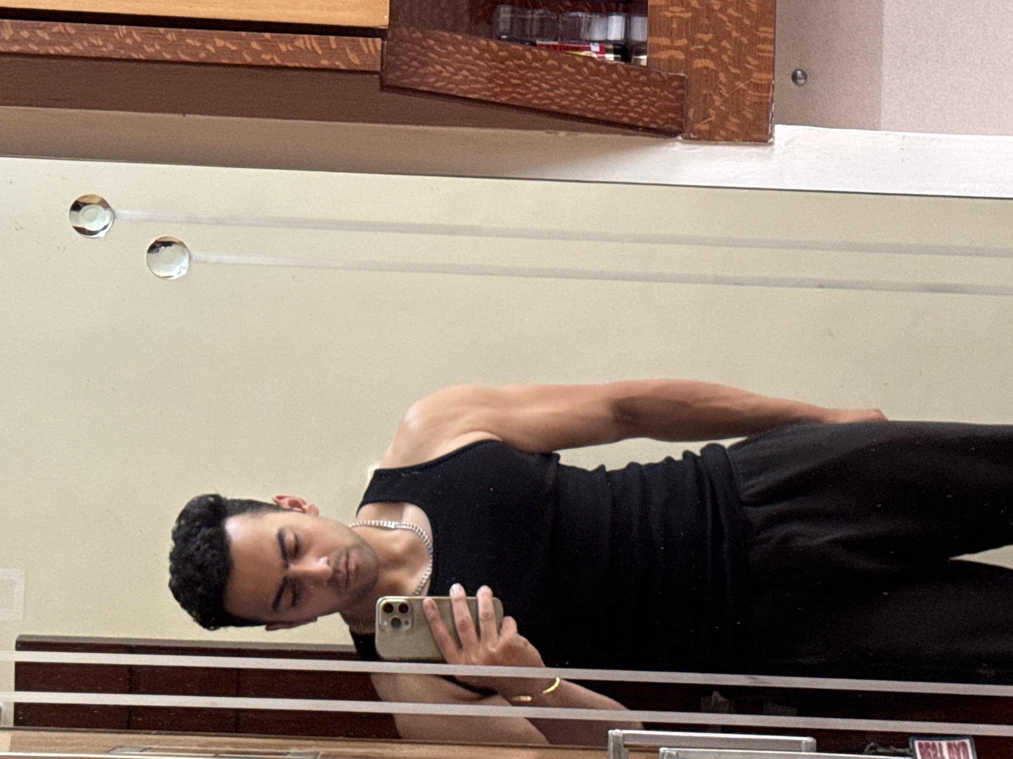 A man is reflected in a mirror. He is wearing a black tank top and black pants. He is holding a mobile phone in his right hand. The mirror is attached to a wall. The location is indeterminate and there is no evidence to support Kolkata, India.
