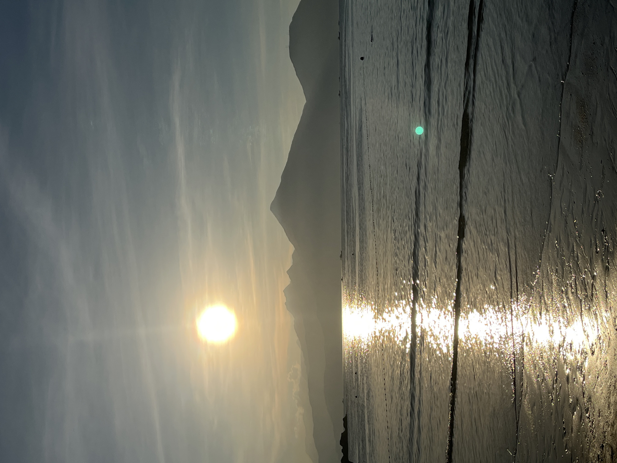 The image, originally captured in a landscape orientation but presented rotated 90 degrees clockwise, portrays a tranquil coastal scene during either sunrise or sunset in Đà Nẵng, Vietnam. The sun, a brilliant golden orb, is positioned low on the left side of the frame, casting a bright glow across the sky and creating a dazzling, shimmering path of light on the water.
The sky in the upper left quadrant is a soft grey-blue with faint, wispy clouds. Along the left half of the horizon, a range of mountains or hills rises as dark, hazy silhouettes against the radiant sky, their edges softened by atmospheric haze.
To the right, a wide expanse of water reflects the sun's intensity, with sparkling highlights dancing on the gentle ripples and waves. In the upper-right section of the water, a small, dark, circular object, likely a buoy, is visible, and a faint, dotted line extends from it towards the foreground, possibly indicating a fishing net or markers. Two distinct, bright green circular lens flares also appear in the water. The immediate foreground on the right reveals wet sand or shallow water, textured with linear patterns from receding waves, further reflecting the golden light. The scene evokes a sense of peace and natural beauty at the transition of day. No text is visible.
