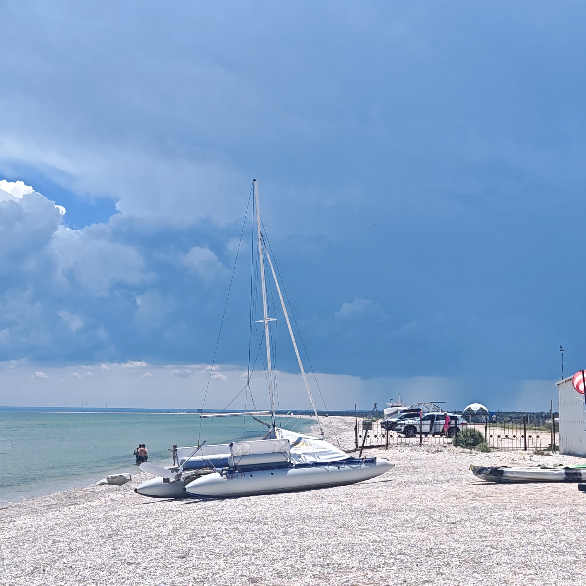 This image depicts a beach scene with a catamaran sailboat grounded on the shore. The beach is covered in small white pebbles. In the background, the ocean meets the sky under a dramatic, cloudy sky with patches of blue. Several people are visible near the water's edge, some wading in the shallow water. There are also several vehicles and a fence visible further up on the beach, suggesting a developed area nearby. A white building with a red and white sign is on the far right. Two kayaks are also visible on the beach in the foreground.