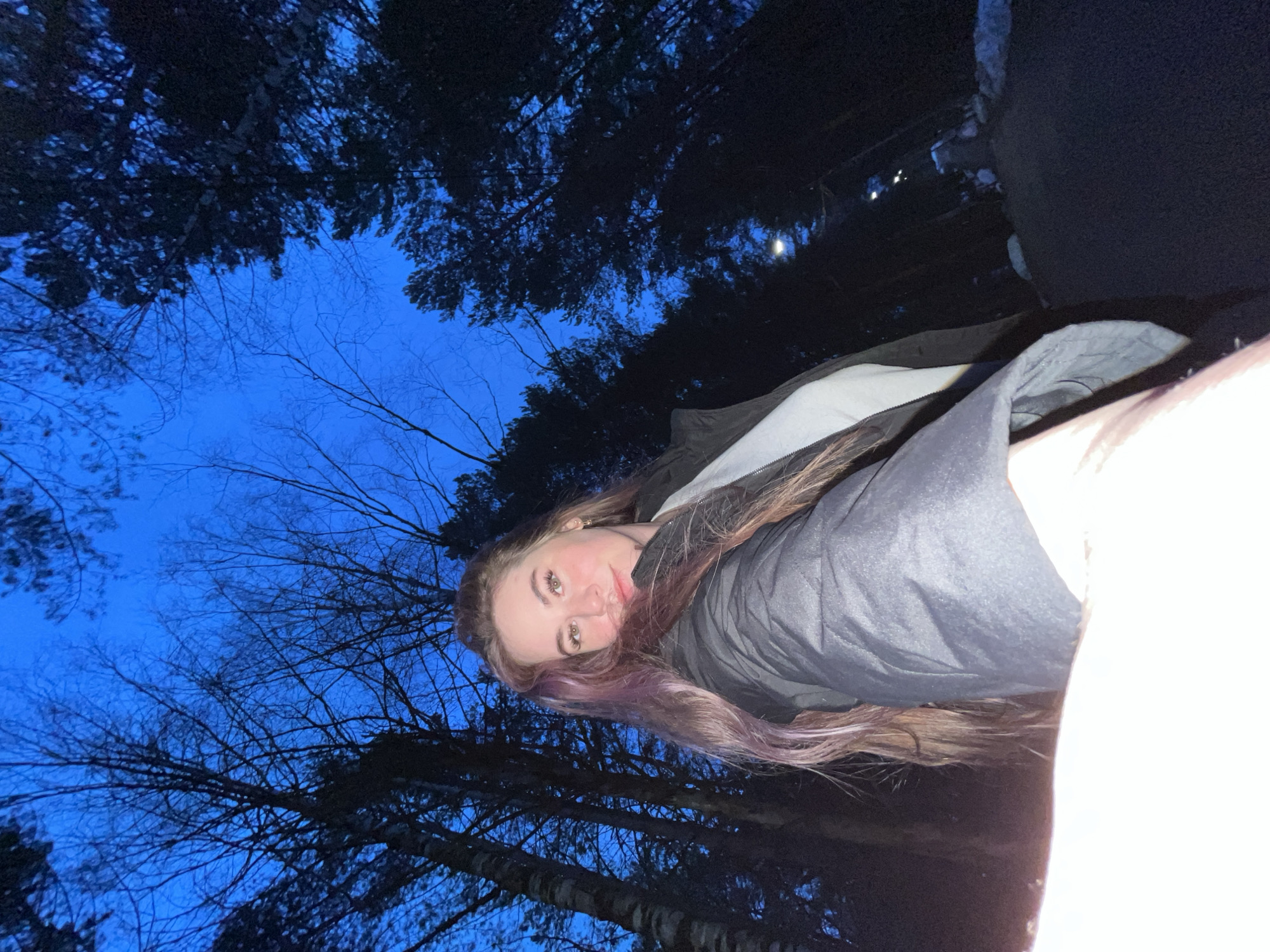 A selfie photo of a woman with brown and light purple hair, wearing a gray jacket. The background is filled with trees and a blue sky, suggesting it is taken outdoors, possibly in a forest or park area. The photo is taken at night. It is not possible to identify the location of the city of Chaikovsky, Russia.