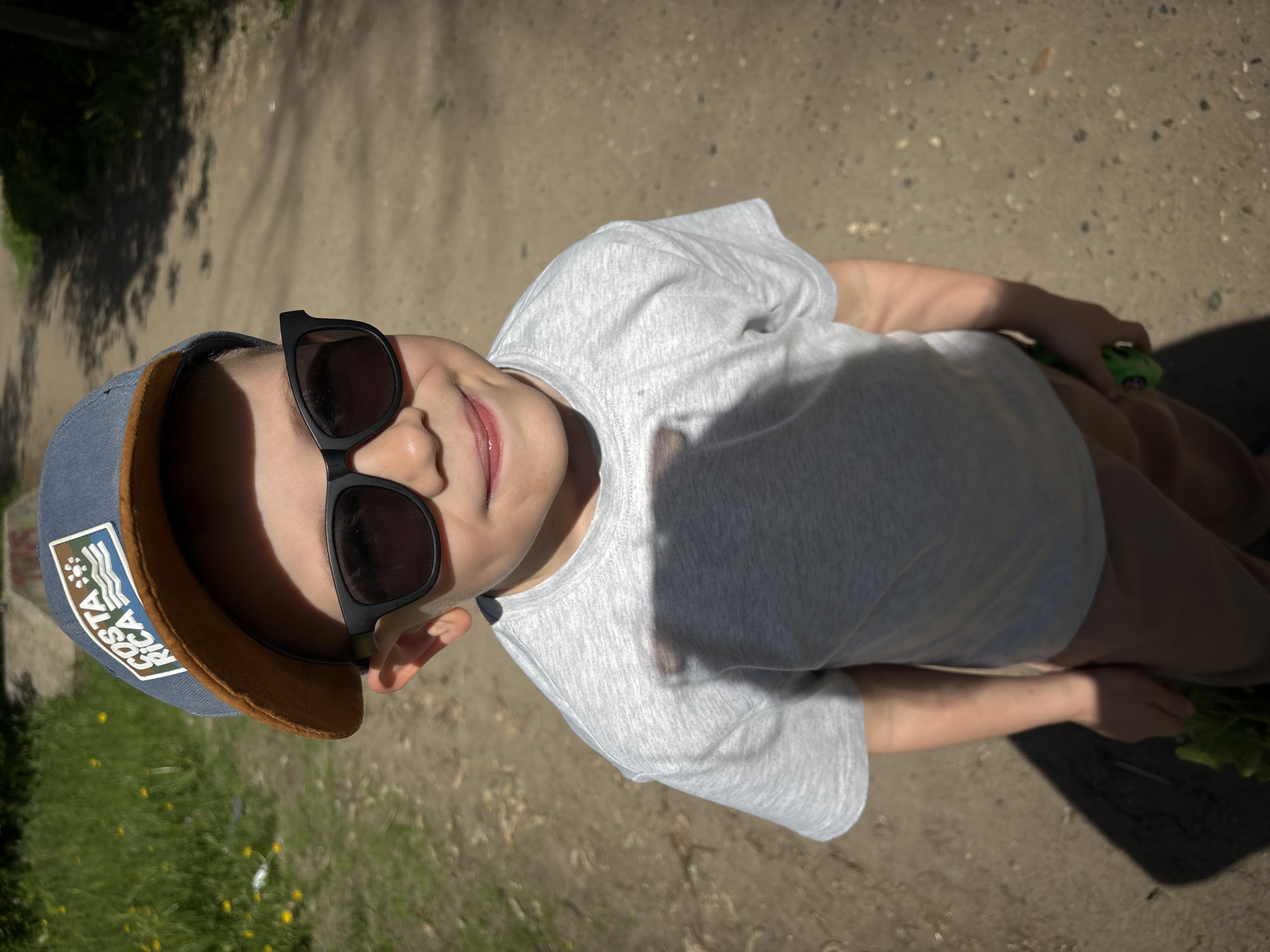 A child wearing a gray t-shirt, a blue and brown baseball cap with a "COSTA RICA" patch, and black sunglasses is seen from a high angle, looking upwards. The child is holding a green object (possibly a toy or plant) in their right hand. The background consists of a dirt or gravel surface, possibly a path or road. The image appears to have been taken outdoors in daylight, with some areas in shadow. There is no discernible evidence to confirm this image was taken in Bolkovo, Russia.