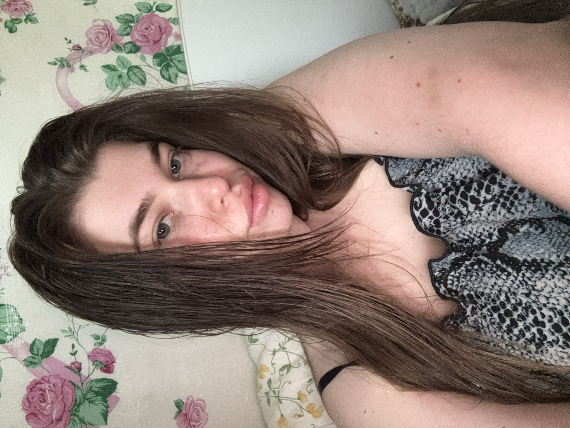 A woman with long brown hair, fair skin, and blue eyes is lying down. She is wearing a printed top with a ruffled neckline. The background features a floral patterned wallpaper and a floral patterned pillow. There is no indication in the image as to its location in the city of Мещёры, Russia, or any other specific location.