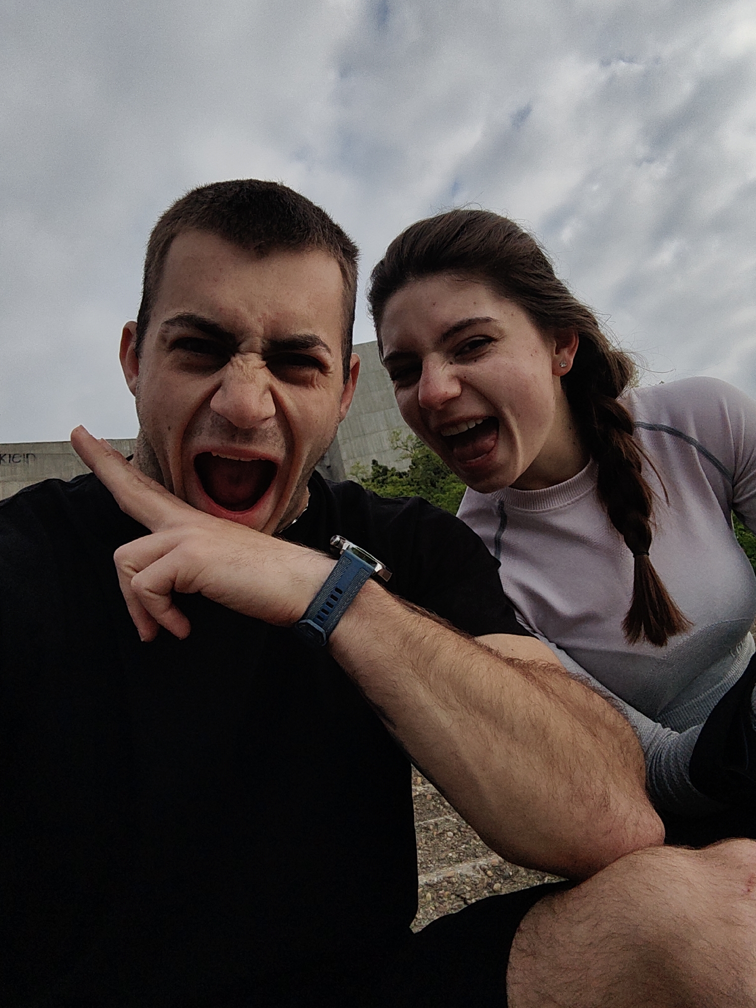 Two individuals are taking a selfie outdoors, likely in Varna, Bulgaria. A man is in the foreground wearing a black t-shirt and a blue smartwatch. He is holding his arm out with a pointed finger. A woman is behind him, wearing a long-sleeved light-colored shirt and a braid. Both are making exaggerated facial expressions with their mouths open.
In the background, a concrete building with the faded word "KLEIN" is visible. The sky is overcast with white clouds. The surface they are sitting on appears to be stone steps or concrete.