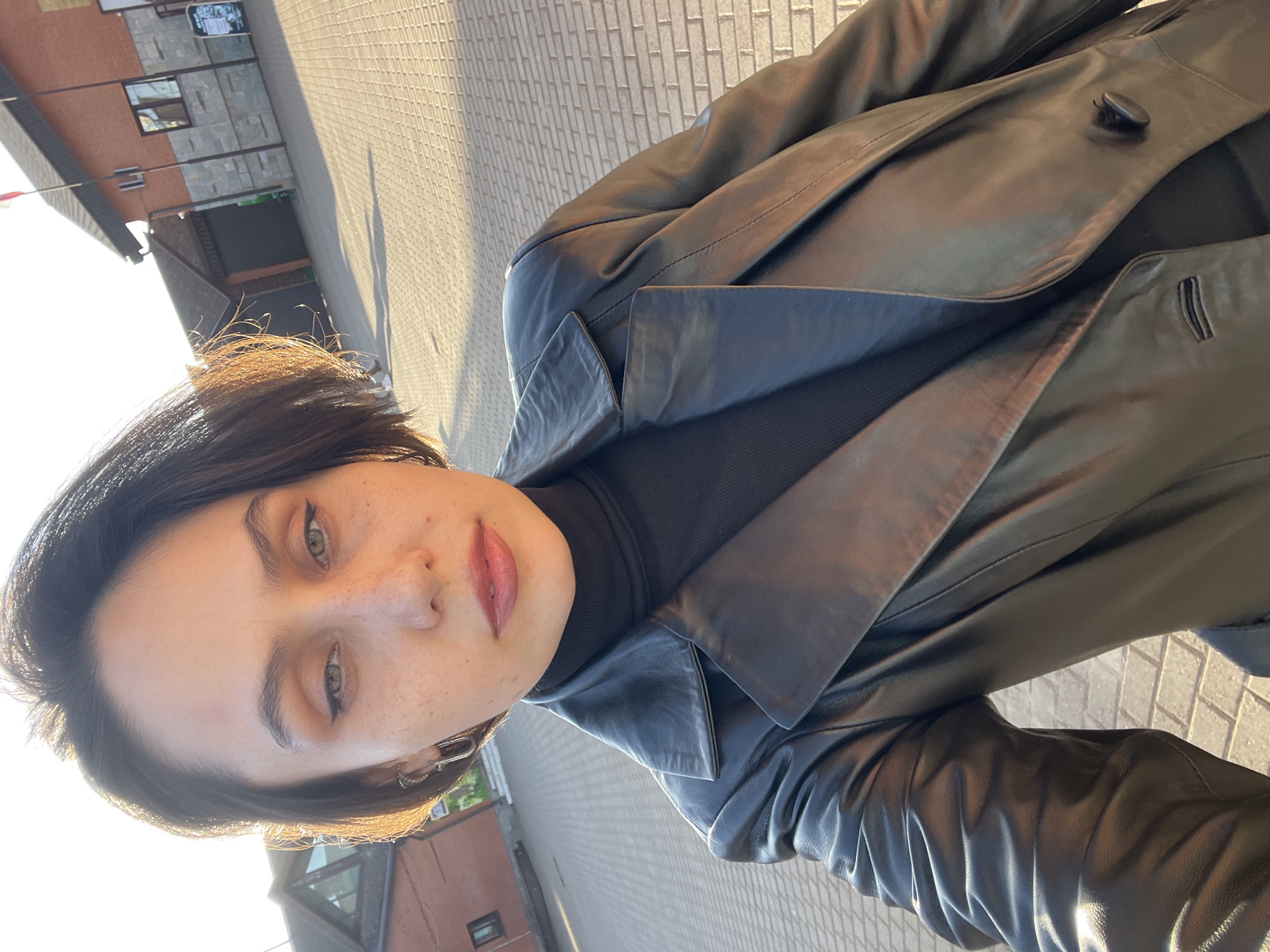 A woman is taking a selfie in an urban area. She is wearing a black turtleneck sweater and a long, dark leather jacket. The background shows a building with windows and a gray brick pavement. The light suggests it may be sunset or sunrise. The location appears to be Izhevsk, Russia based on contextual clues.