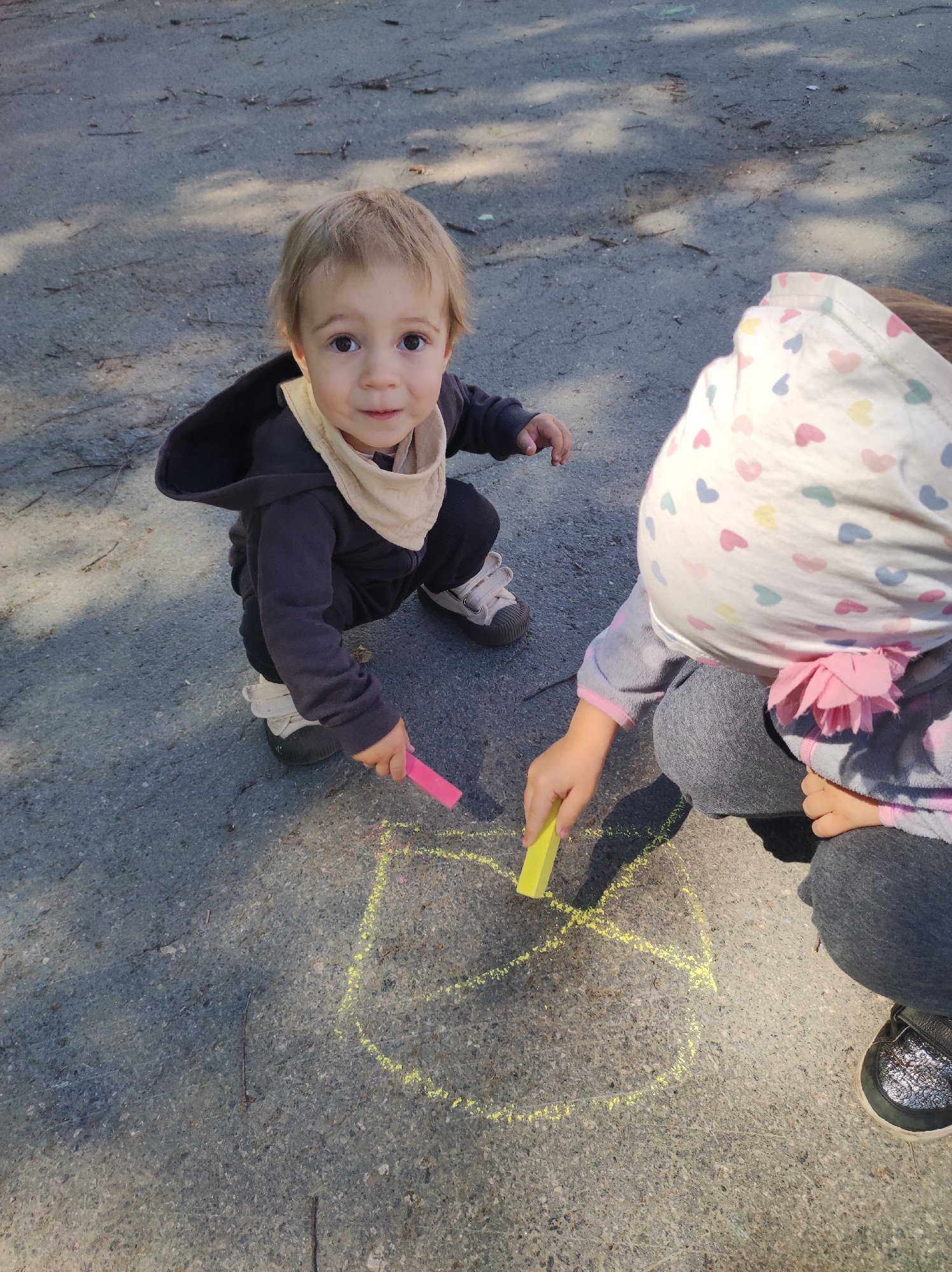 Two children are drawing with chalk on an asphalt surface. One child, with light blonde hair, is crouching and looking directly at the camera while holding a pink chalk. This child is wearing a dark gray hooded jacket, dark pants, and light-colored shoes. The second child, who is closer to the right side of the frame, is wearing a head covering patterned with hearts, a gray long-sleeved shirt with pink trim, and dark pants. This child is holding a yellow chalk and drawing on the asphalt. A yellow chalk drawing is visible on the ground. The asphalt surface is light gray with small bits of debris and shadows. The location in Chaikovsky, Russia, is not discernible from the image.