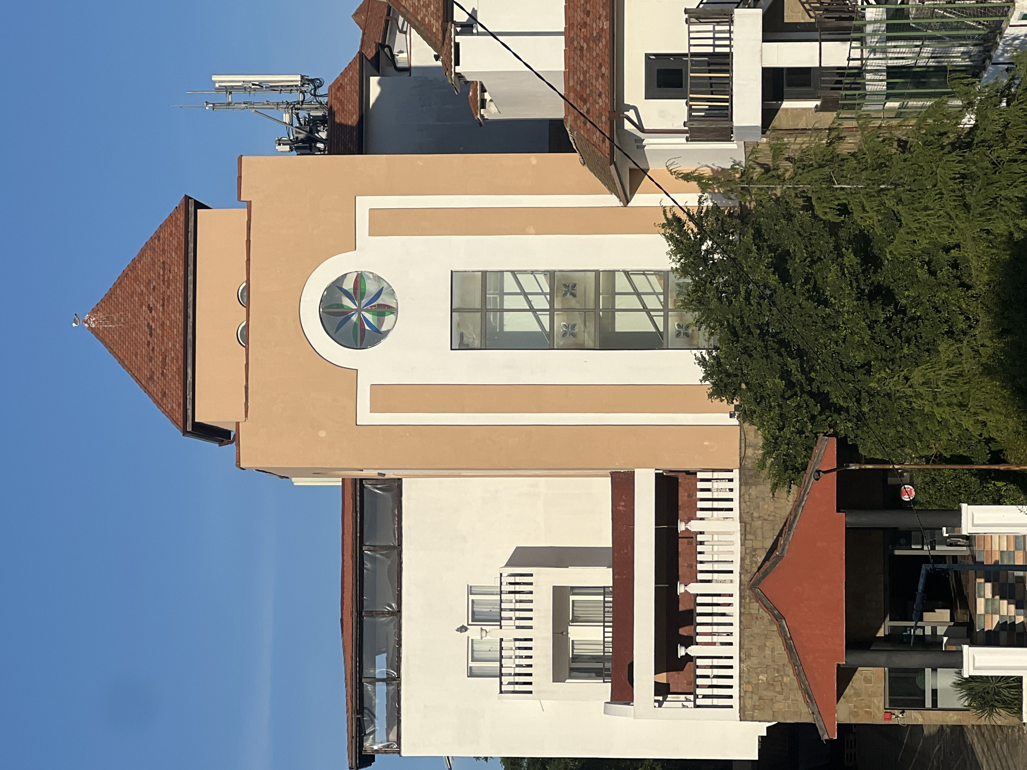 This is a full outdoor, eye-level, vertical shot of a multi-story building with a reddish-brown tiled roof and pale orange and white painted walls. The building features several balconies with white or dark wooden railings, multiple windows, and a decorative stained-glass circular window with a colorful geometric pattern. A telecommunications tower is visible on the roof of a section of the building. The building is surrounded by lush green trees and foliage. The sky is clear and blue. The architectural style suggests a modern resort or hotel, possibly in a coastal area given the potential for similar structures in Sinemorets, Bulgaria, a town known for its beaches and natural beauty. No people are visible in the image.