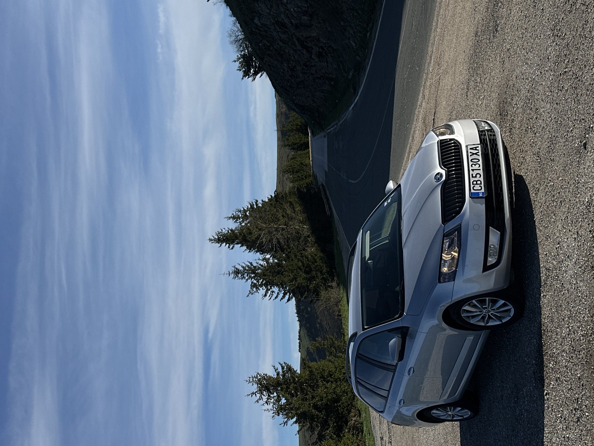 A silver Skoda Octavia Combi is parked on a paved road, the front of the vehicle facing slightly upward in the frame. The license plate reads "CB 5130 XA." The background includes a road winding along a hillside covered in trees. The sky is blue with streaky white clouds. The location is near Hristo Danovo, Bulgaria, based on the vehicle registration. The lighting suggests it is daytime. No people are visible in the image.