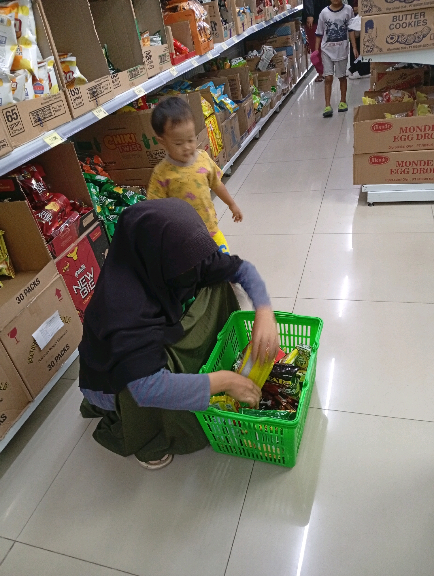 The image depicts an indoor scene, likely a supermarket aisle in Babakan, Indonesia. In the foreground, a person wearing a black hijab and an olive green outfit is crouching down, rummaging through a bright green plastic basket filled with various food items, some of which appear to be snacks or packaged goods. The person's hands are visible as they select an item. To the left, part of a cardboard box with "30 PACKS" printed on it is visible. In the mid-ground, a young child in a yellow shirt with a graphic print is standing and looking towards the basket. Behind the child, a shelf displays more packaged goods. Further back, a young boy in a white t-shirt and grey shorts is walking away from the camera. The aisle has light-colored tiled flooring and shelves stocked with a variety of products, including boxes and bags of snacks and other groceries. The overall environment suggests a retail setting.