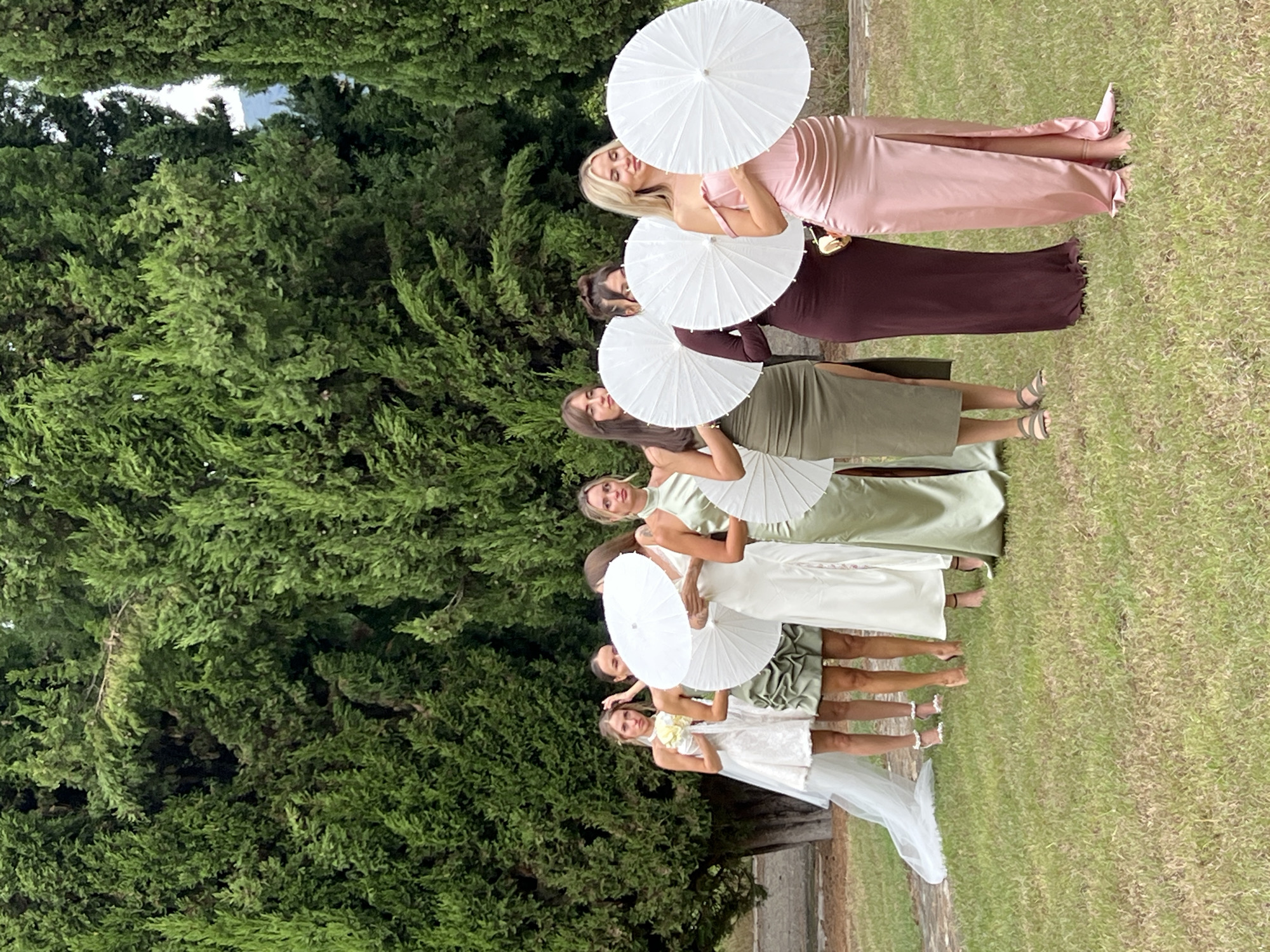 An outdoor, full shot captures a procession of seven women dressed in formal attire, standing in a single file line against a backdrop of lush green cypress trees. The woman at the front, on the left, is wearing a short white lace wedding dress with a veil. The other six women are dressed in floor-length gowns in shades of sage green, dark plum, and rose pink, each holding a white parasol. The ground is covered in a well-maintained lawn. The scene appears to be a wedding party posing for a photograph, possibly in a garden or park setting. There are no visible indications of the specific location in Saniore, Georgia.