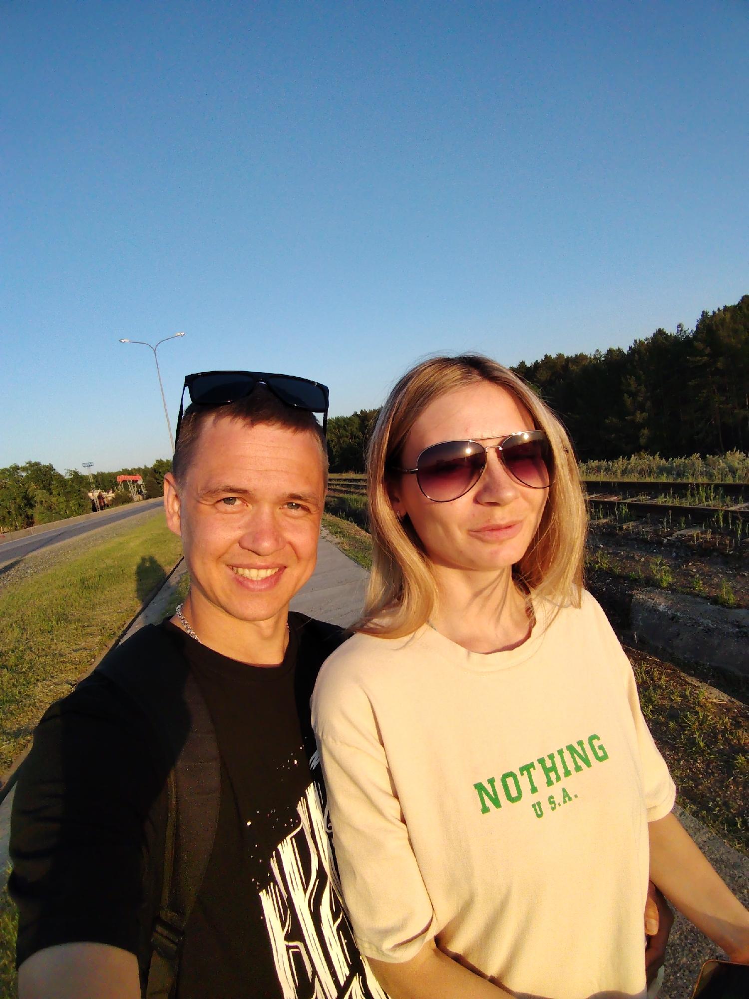 A selfie-style photograph shows a man and woman standing close together in what appears to be an urban or suburban environment. The location is Chaikovsky, Russia. The man is on the left, smiling, wearing a black t-shirt with a white design on the front. He has sunglasses resting on his head and a silver chain around his neck. The woman is on the right, wearing oversized sunglasses and a light yellow t-shirt with the word "NOTHING" printed in green across the chest. Behind them is a grassy area with a road to the left, trees and railroad tracks to the right. The sky is clear and blue. A lamppost is visible on the road. The photograph appears to be taken in daylight.
