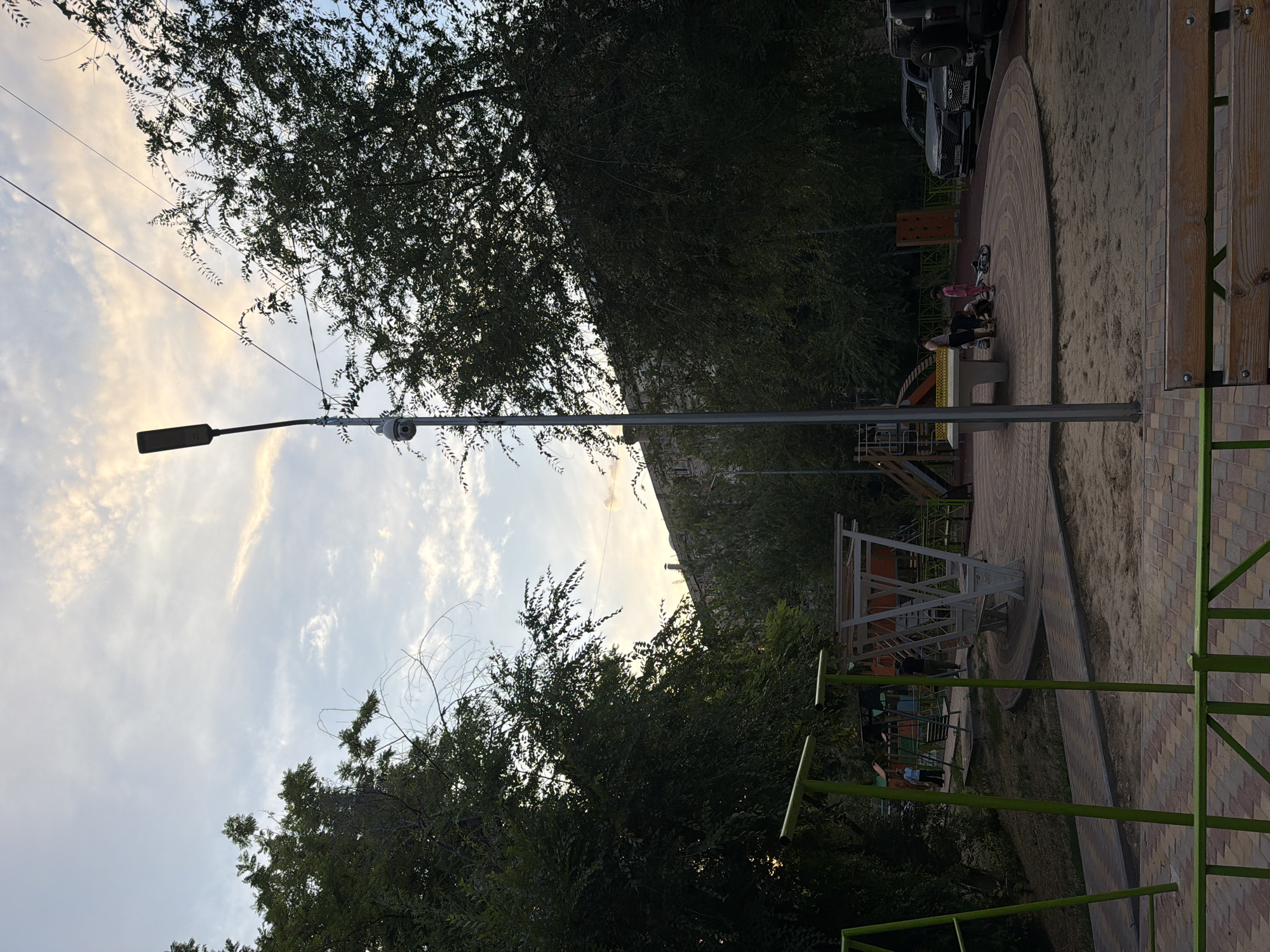 The image depicts an outdoor children's playground in the city of Shymkent, Kazakhstan. The playground features a sandpit, a table tennis table, swings, and a slide. Several children and adults are present in the playground, some of whom appear to be playing. The playground is surrounded by trees and a building, suggesting an urban setting. A streetlight with a surveillance camera is visible in the foreground. The sky is partly cloudy, with the sun setting in the background. The ground is paved with colorful bricks and sand. Cars are parked in the background, near the building.