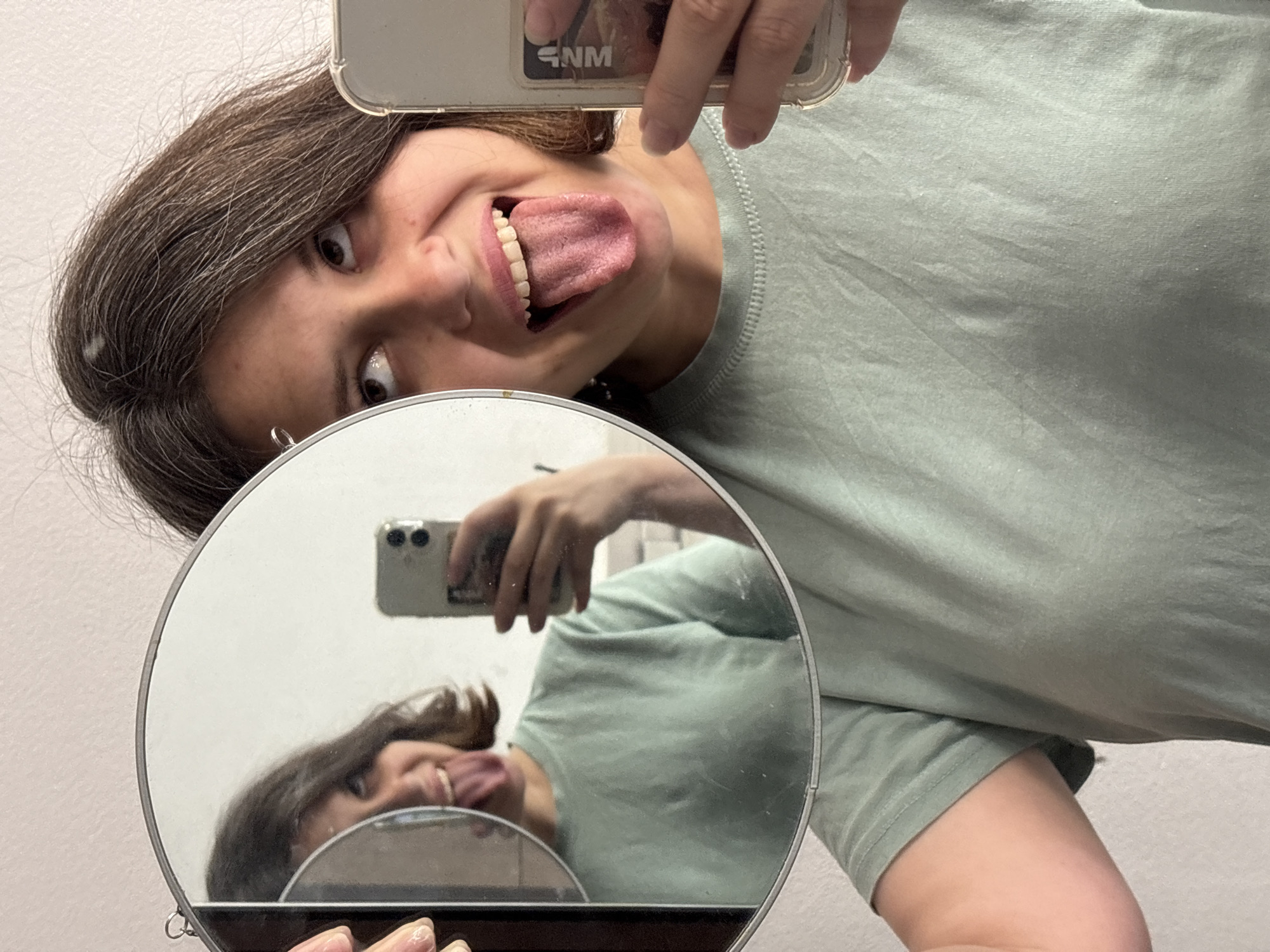 A woman is holding a mirror to take a selfie while sticking her tongue out. She is holding a phone in her left hand to take the picture. The background is a plain wall. It is not possible to determine if this picture was taken in Chaikovsky, Russia.