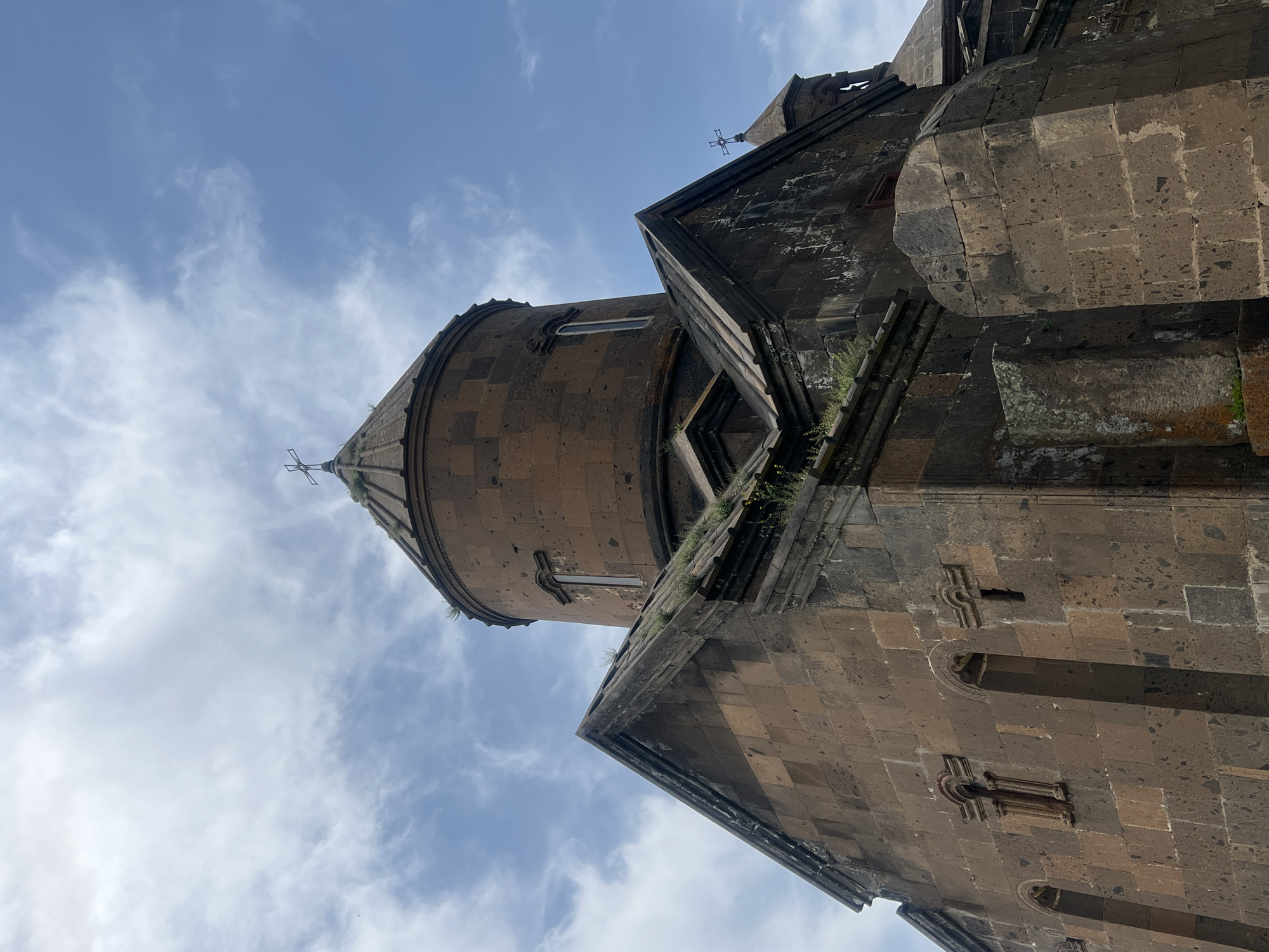 The image shows a low-angle view of Saghmosavank Monastery in Artashavan, Armenia. The architecture is dominated by dark stone construction against a backdrop of a cloudy blue sky. The dome and roofline of the religious structure are visible, featuring a cross at the apex. Patches of vegetation are growing on the structure's surface.