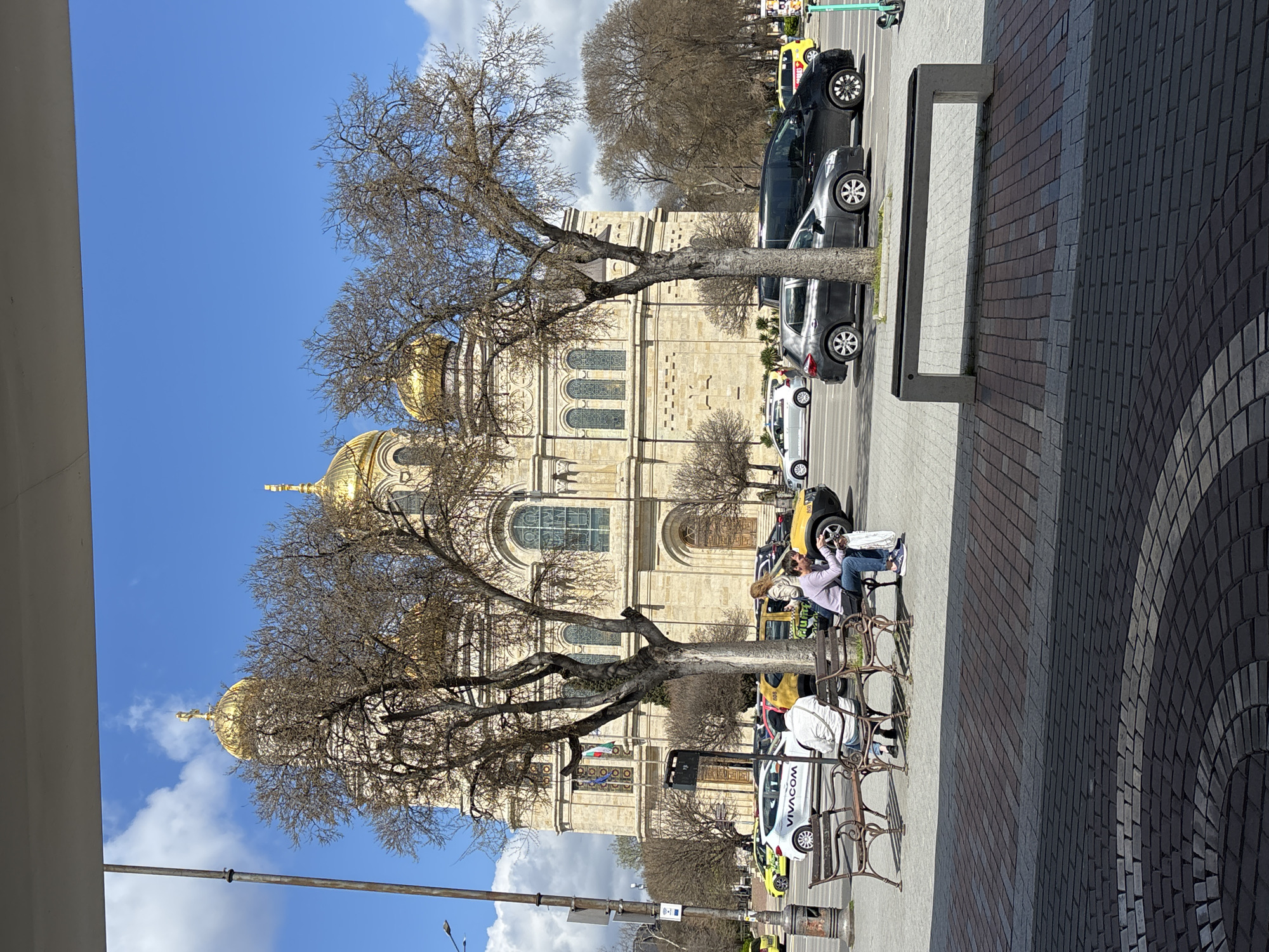 The image shows a daytime scene in Varna, Bulgaria, featuring the Dormition of the Mother of God Cathedral. The cathedral, with its golden domes, is partially obscured by bare trees. Cars are parked along the street in front of the cathedral. People are sitting on benches. The sky is blue with some clouds. The foreground includes a sidewalk with a patterned brick surface. There's a white support column on the left. A street lamp is visible.