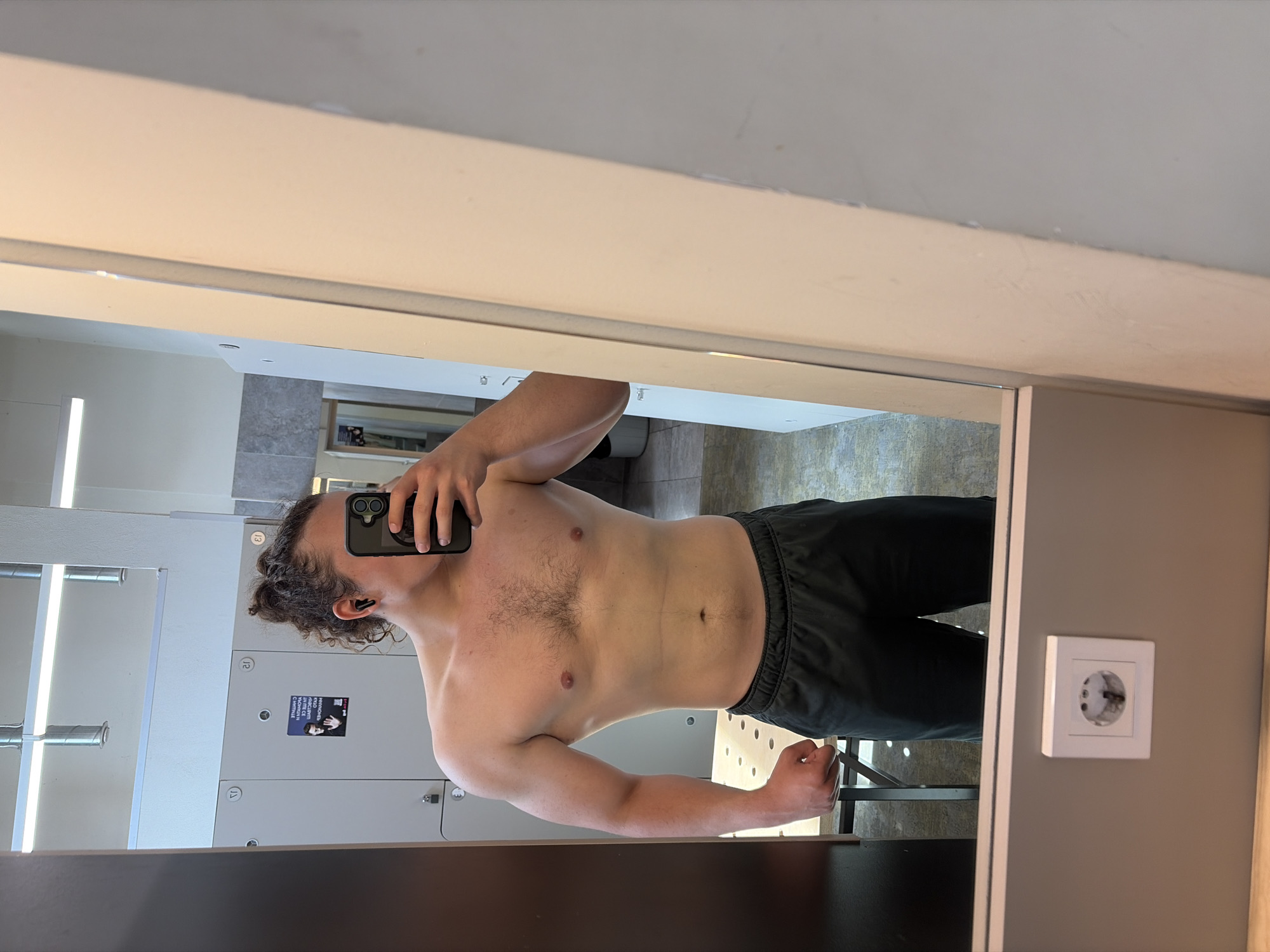 A man is taking a selfie in what appears to be a gym changing room. He is shirtless and wearing dark-colored athletic pants. The room has lockers on the left, a mirror in front, and a power outlet on the wall to the right of the mirror. The reflection in the mirror shows a glimpse of a window or another room. There is a poster on one of the lockers. The man has curly hair and is holding a black smartphone up to take the picture. There is no verifiable information to confirm a specific location, such as Plovdiv, Bulgaria.
