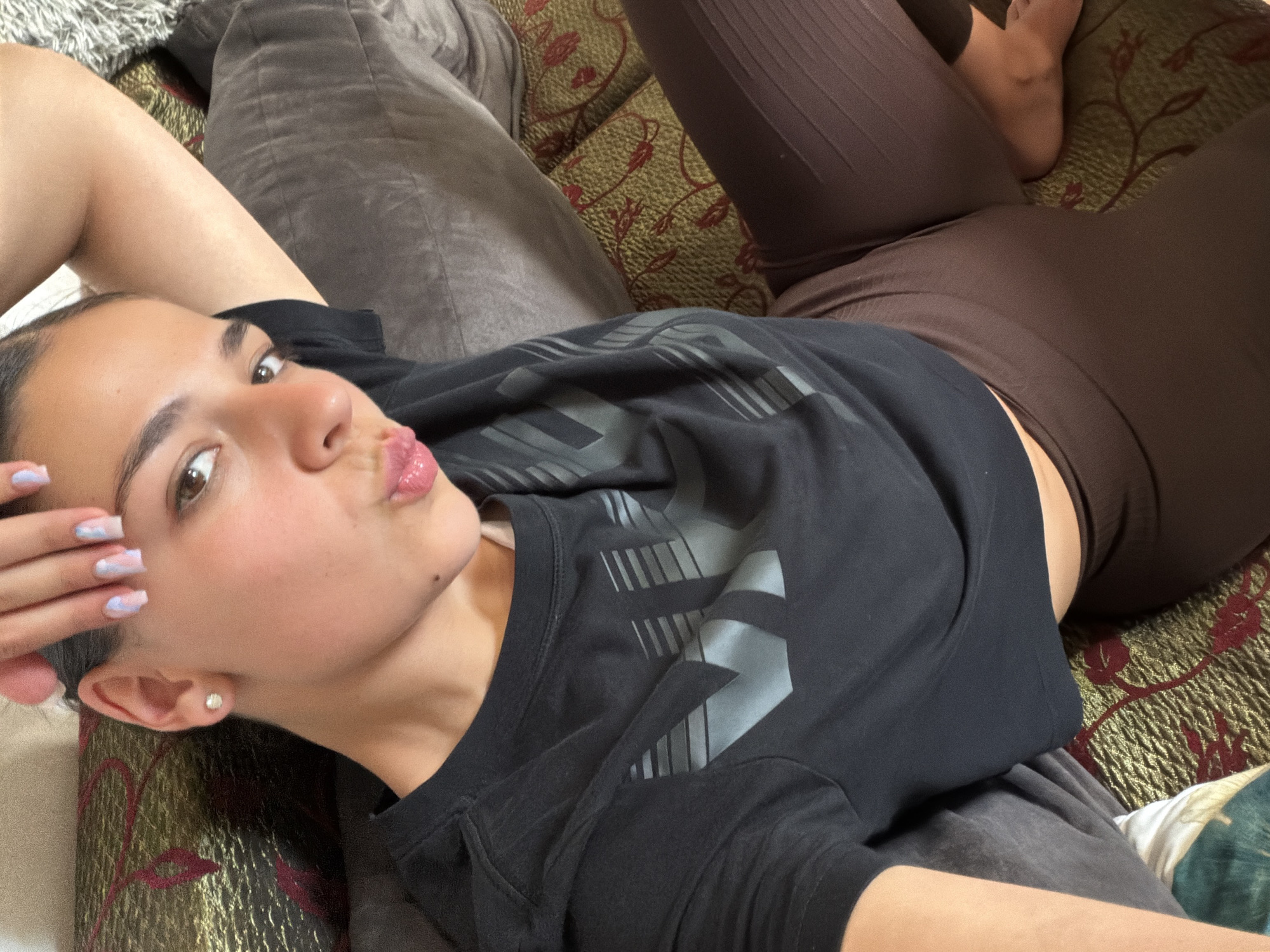 A young woman lies on a patterned couch. She is wearing a black shirt and brown leggings. She has light skin and brown eyes, and her lips are pursed in a kiss. Her left hand is raised near her face, and her nails are painted with a light blue and white design. She is indoors, and the background is out of focus. No information suggests the location is Gabrovo, Bulgaria.