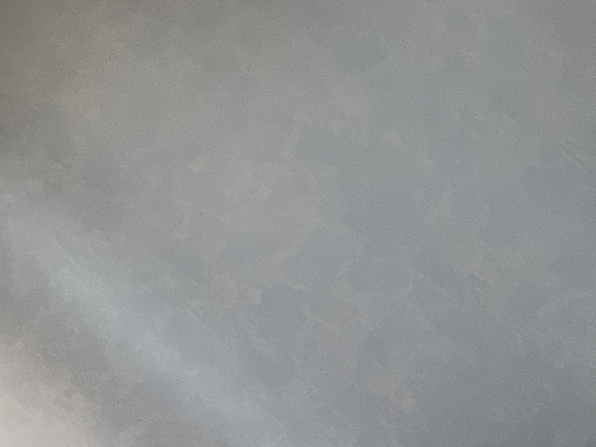A close-up, full-frame, vertical, overhead shot of a textured gray surface is presented. The texture is subtle and appears to be a fine-grained stucco or concrete. The lighting is soft and diffuse, creating subtle variations in tone across the surface, with lighter areas in the upper left quadrant and darker, more uniform gray throughout the rest of the image. There are no discernible objects, people, or actions depicted in the image. The background is entirely this textured gray surface.