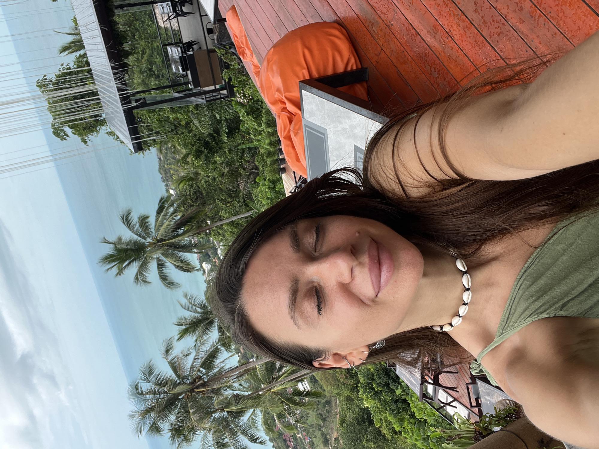 A medium, upward-angle shot shows a woman with tan skin and brown hair wearing a green top and a shell necklace, with her eyes closed, appearing serene. The background reveals an outdoor setting, likely a resort or elevated area, featuring palm trees and a view of the ocean. An orange beanbag-style seat and a wooden deck suggest a relaxation area. There is a building structure with glass panels visible in the background. The location appears to be a tropical environment. The city and location are unknown.