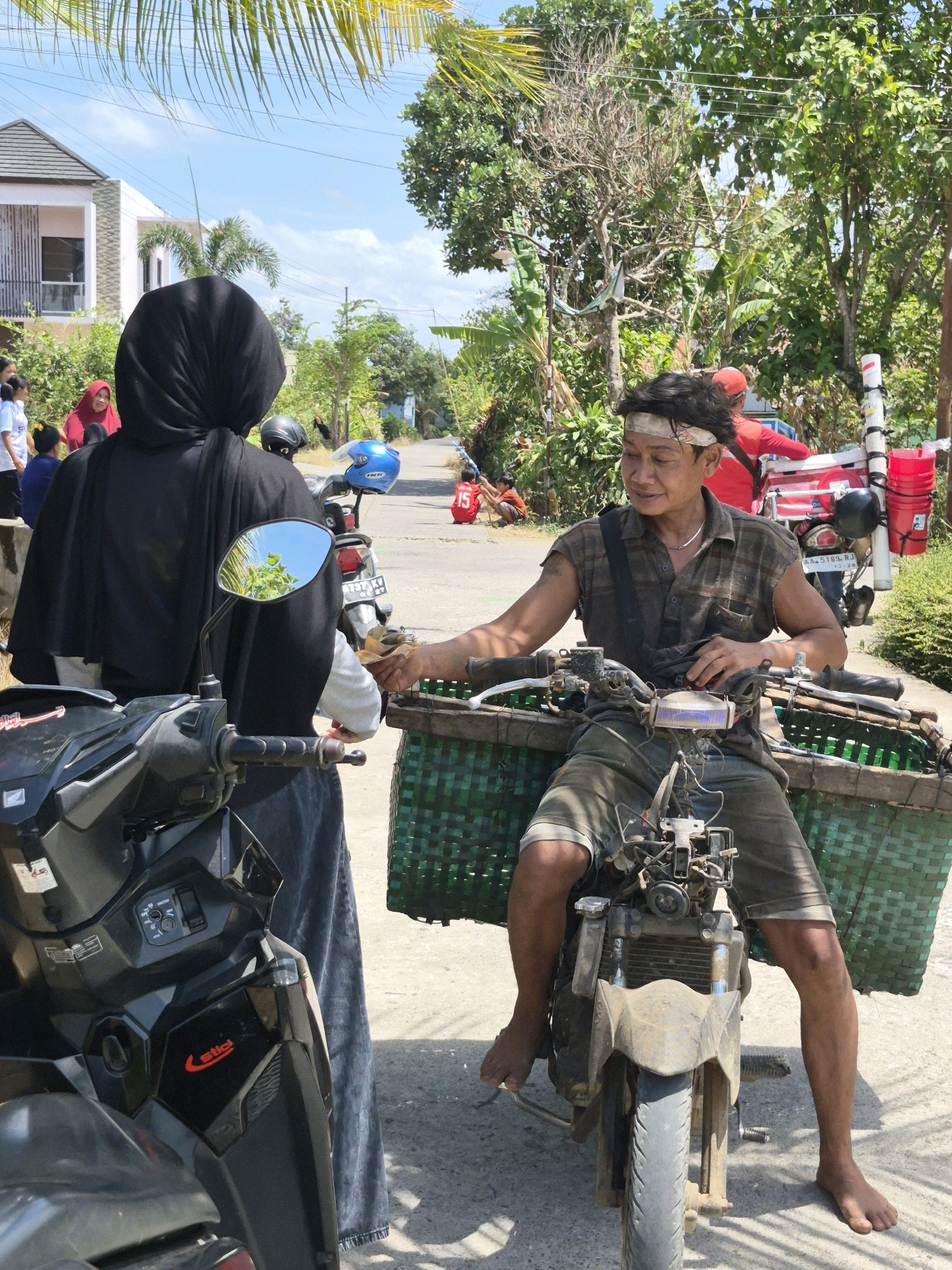 In Grantung, Indonesia, a man sits on a motorcycle with a large green woven basket attached to the front. He is wearing a plaid sleeveless shirt, shorts, and a headband, and is handing something to a person standing beside him, who is wearing a black hijab and a blue skirt. The motorcycle is dirty, with visible mechanical parts. The background shows a paved road lined with trees and houses, suggesting a residential or semi-rural area. The sky is bright blue with some clouds. There are other people and vehicles visible in the distance.