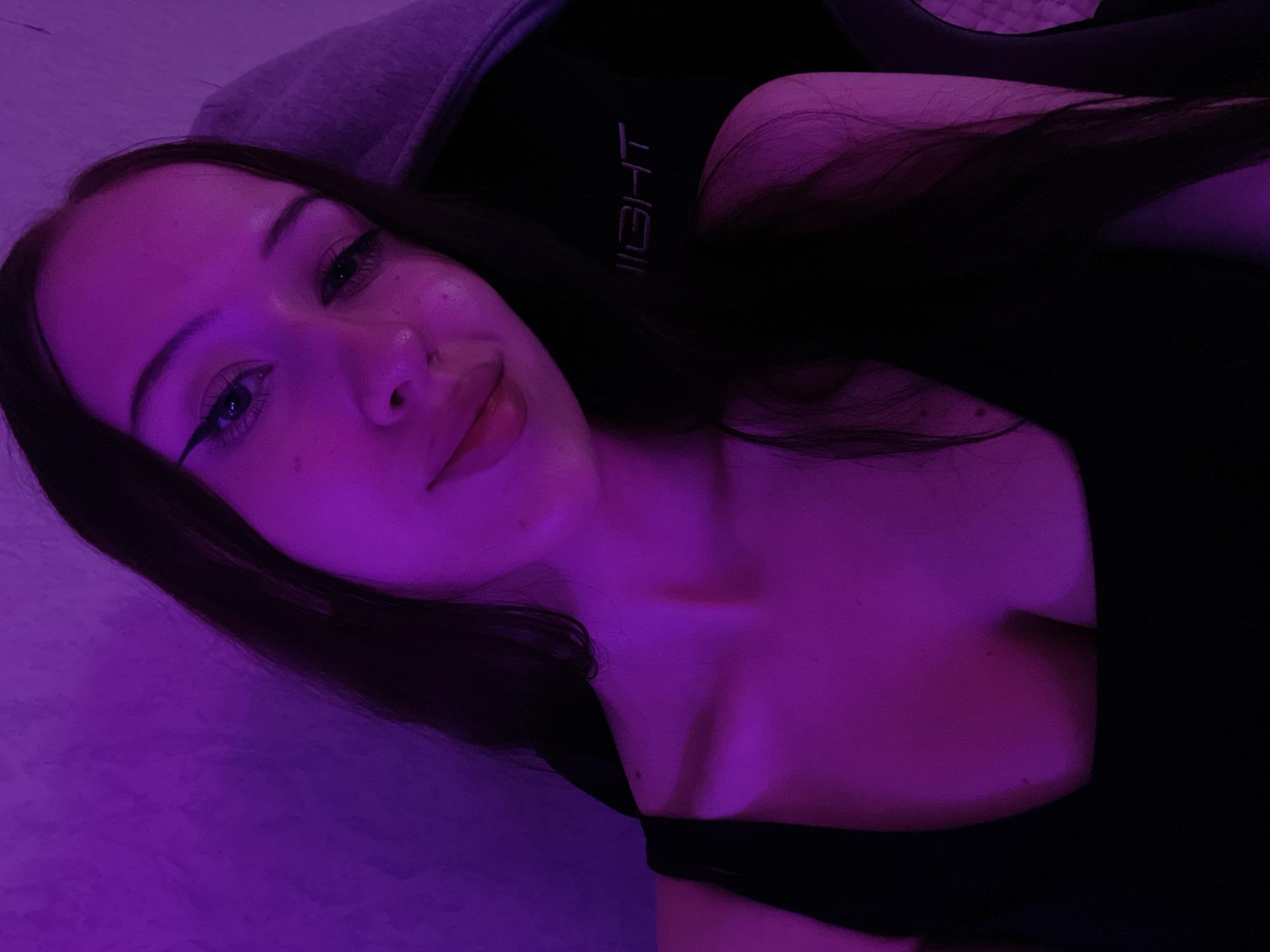 The image is a selfie of a young woman with long, dark hair, wearing a black tank top. She is illuminated by purple lighting, which casts a vibrant glow on her face and upper body. Her makeup includes winged eyeliner. The background appears to be a wall with a textured surface. A portion of a gray hooded garment is visible behind her right shoulder, and a black chair with the embroidered word "LIGHT" is behind her left shoulder. The scene does not contain any information that allows for localization in Votkinsk, Russia.