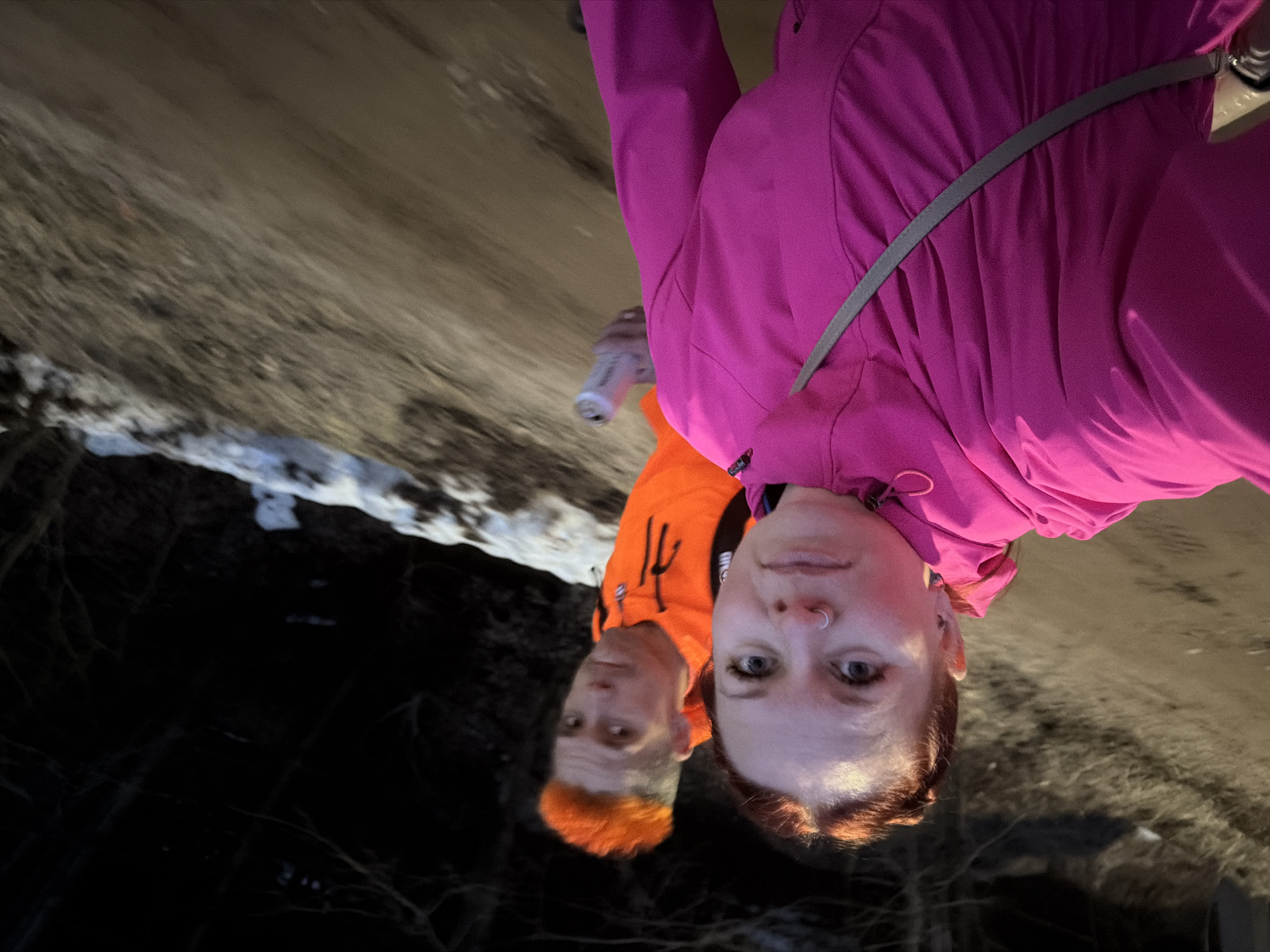 Two individuals are visible in a selfie-style image. The individual in the foreground wears a bright pink jacket with a grey strap crossing the chest. The person behind is wearing a bright orange jacket. The background consists of a mixture of dark areas and possibly snow or light-colored groundcover. The photo was probably taken in Chaikovsky, Russia, although there is no evidence of it in the image.