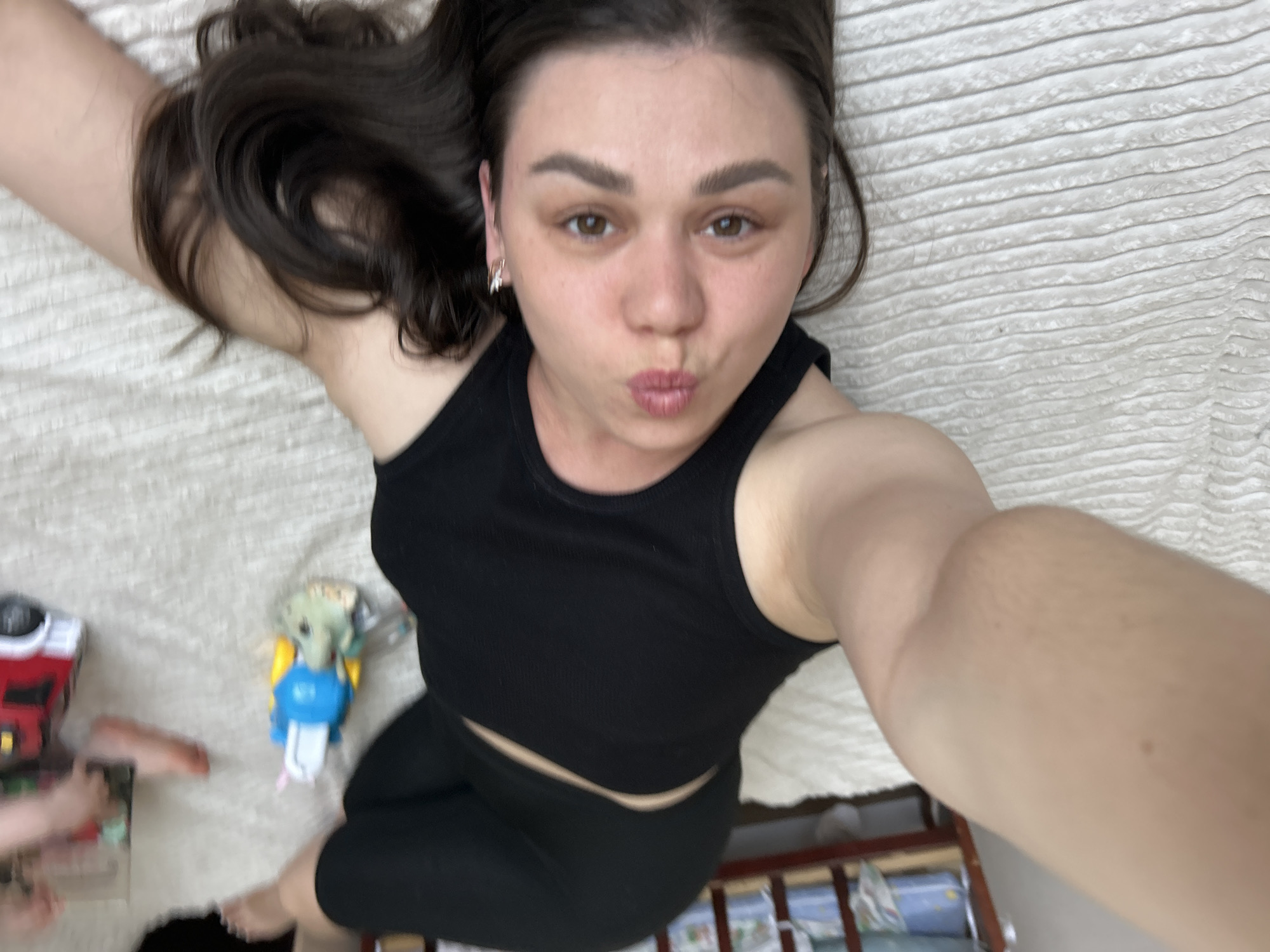 A person, presumably female, is lying on a bed taking a selfie. She is wearing a black tank top and black leggings. Her dark hair is spread out on the white bedspread. She has brown eyes and is making a kissing face. To the side of the person are toys and a baby crib. The location is presumed to be the city of Chaikovsky, Russia.