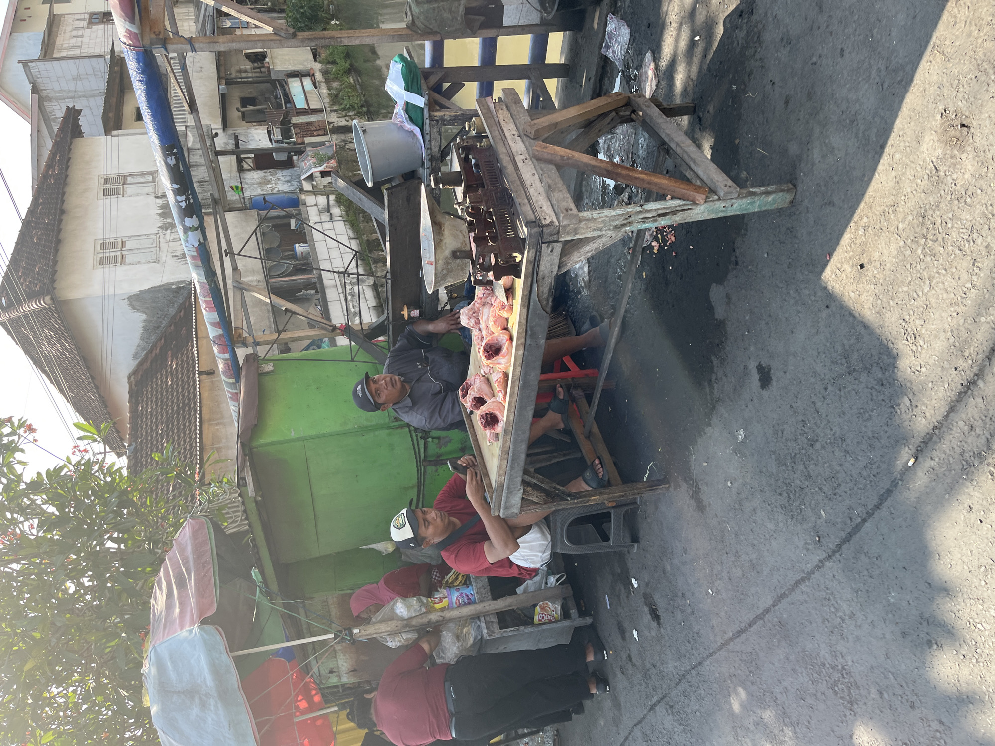 In Tulangan, Indonesia, on a street, two men are engaged in selling raw chicken. One man, wearing a red t-shirt and white shorts, sits on a black stool, holding a smartphone. The other man, in a grey shirt and black cap, sits on a red stool, looking to his left. Both are positioned in front of a wooden table displaying several cut chicken carcasses. To their left, a stall with a green facade is visible, featuring two women in red and pink attire. The scene unfolds outdoors, with buildings in the background and a body of water to the right. The ground is paved with asphalt, and scattered debris is present.