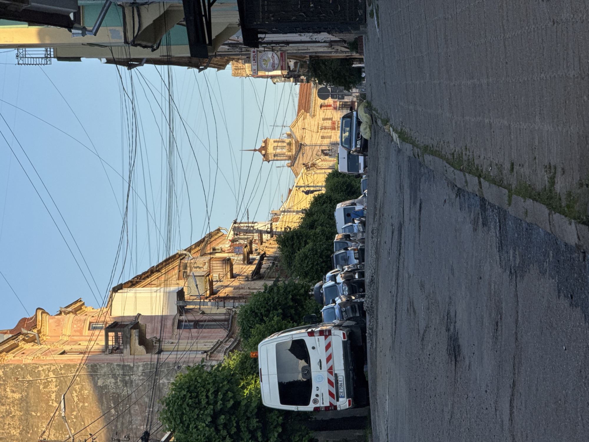 This is a vertically oriented, eye-level shot of a street in Chernivtsi, Ukraine, during what appears to be late afternoon or early evening, given the warm, golden light. The scene depicts a somewhat weathered, historic European street lined with old buildings on the left and a row of parked vehicles on the right.
The buildings on the left display architectural details indicative of age, with peeling paint, ornate cornices, and balconies. A complex network of overhead wires crisscrosses the sky, a common sight in many older urban areas. The buildings are illuminated by the setting sun, casting long shadows and highlighting their textured facades.
On the right side of the street, a line of cars and vans are parked along the curb. The most prominent vehicle in the foreground is a white Mercedes-Benz Sprinter van, clearly marked with red and white reflective stripes on its rear. Other vehicles, mostly dark-colored cars and vans, are visible further down the street. Lush green trees and bushes are interspersed between the vehicles and the buildings, adding a touch of nature to the urban landscape.
The street itself is paved with asphalt, showing signs of wear and tear. The overall impression is one of a lived-in, historic urban environment bathed in the soft light of a fading day. No people are visible in the frame.
Visible text includes "MERCEDES-BENZ SPRINTER" on the back of the van, along with a license plate starting with "CE 7596". A sign above a building on the left reads "ANGELHOLM".