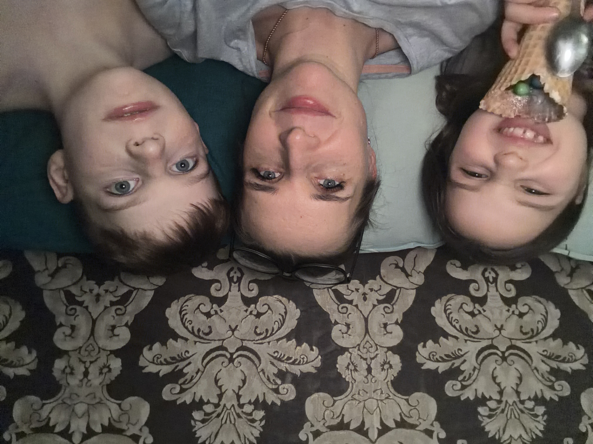 Three individuals, a woman and two children, are positioned together on a patterned surface. The patterned surface is dark brown and gray. The woman is in the center. She is wearing glasses and a light gray shirt with a pink undershirt. To her left is a boy with blue eyes and light brown hair wearing a dark green shirt. To her right is a girl holding an ice cream cone with a spoon. Location: Zaprudnya, Russia.