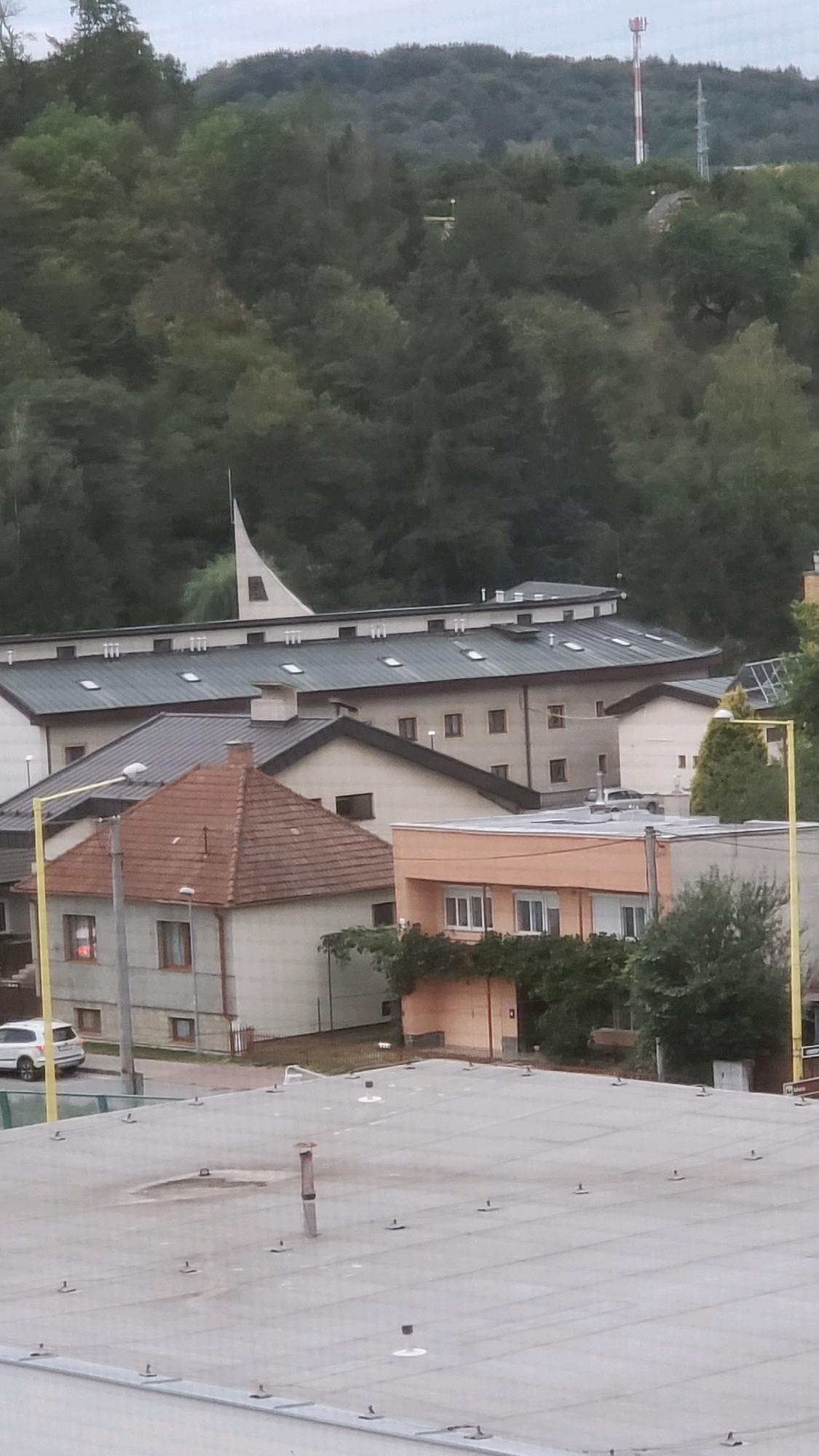 The image shows a residential and institutional area with a backdrop of lush green hills. In the foreground, a flat, grey rooftop with numerous small black anchor-like fixtures and a central ventilation pipe is visible. Beyond this, several buildings are present. One building has a reddish-brown, gabled roof and light-colored walls with windows. Adjacent to it, a larger, multi-story building features a dark, sloping roof with multiple skylights and a light-colored facade with regularly spaced windows. Further back, a distinctive white building with a pointed, steeple-like architectural element rises above the others. In the distance, the forested hills are dotted with trees. A red and white striped tower, likely a communication tower, is situated on the highest point of the hill. A white SUV is parked on the street in front of one of the buildings. The sky is overcast, suggesting cloudy weather. The scene appears to be a daytime view.