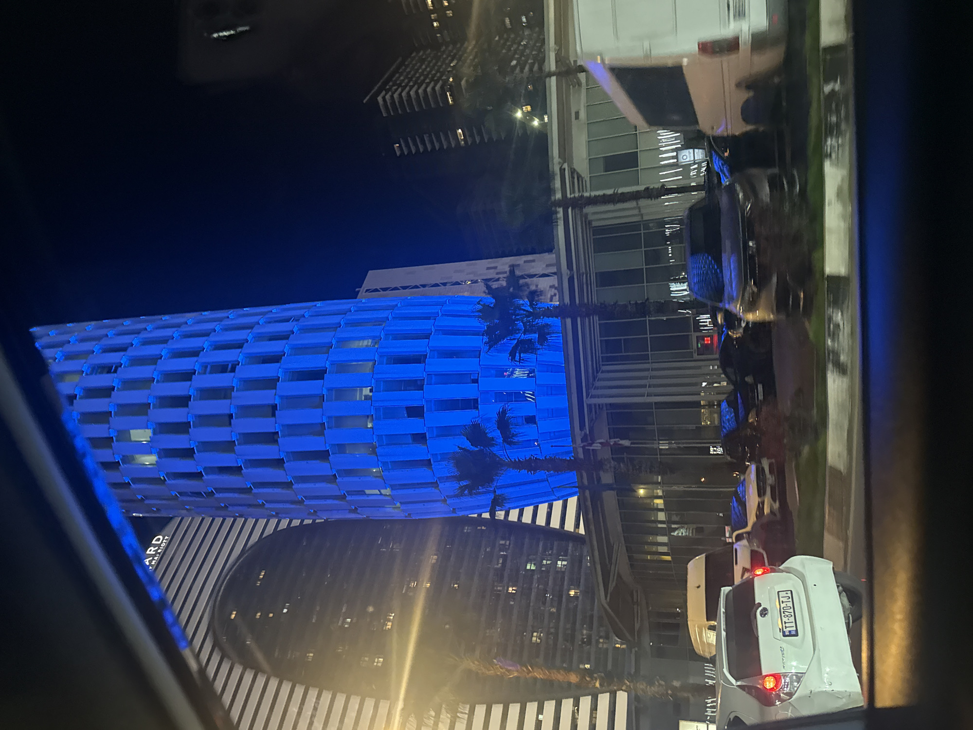Captured from inside a vehicle at night, the image presents a view of illuminated buildings in Batumi, Georgia. The most prominent building is a tall, cylindrical structure lit in blue, featuring a grid-like facade. Next to it, a building with a rounded front and white and gray striped design is visible. Several palm trees are interspersed between the buildings. Streetlights illuminate the road, where several vehicles are parked, including a white car in the foreground. The scene is urban, with a modern architectural style.