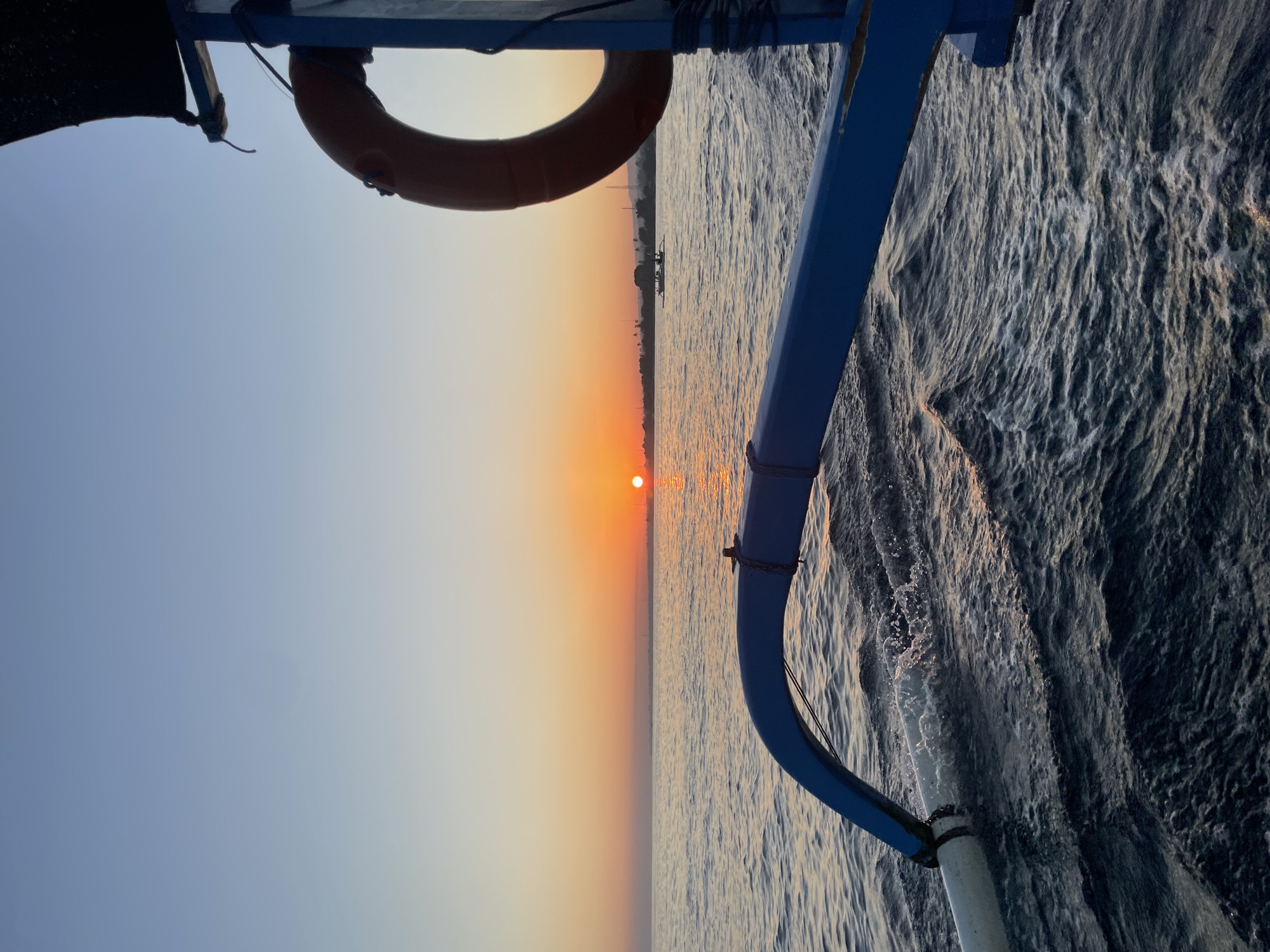 This is a photograph taken from a boat in the ocean near Banjar, Indonesia, at sunrise. The sun is a bright orange orb just above the horizon, casting a warm glow across the sky and water. The sea is calm with gentle waves, and the reflection of the sun shimmers on the surface. In the foreground, a blue outrigger of a traditional Indonesian fishing boat is visible, with a white float attached to its end. Further out, another small boat with people on board can be seen. The coastline with some vegetation and structures is visible in the distance. A life preserver is hanging from the boat's structure on the right side of the frame. The sky is a clear gradient of pale blue at the top, transitioning to a warm orange and yellow near the horizon.