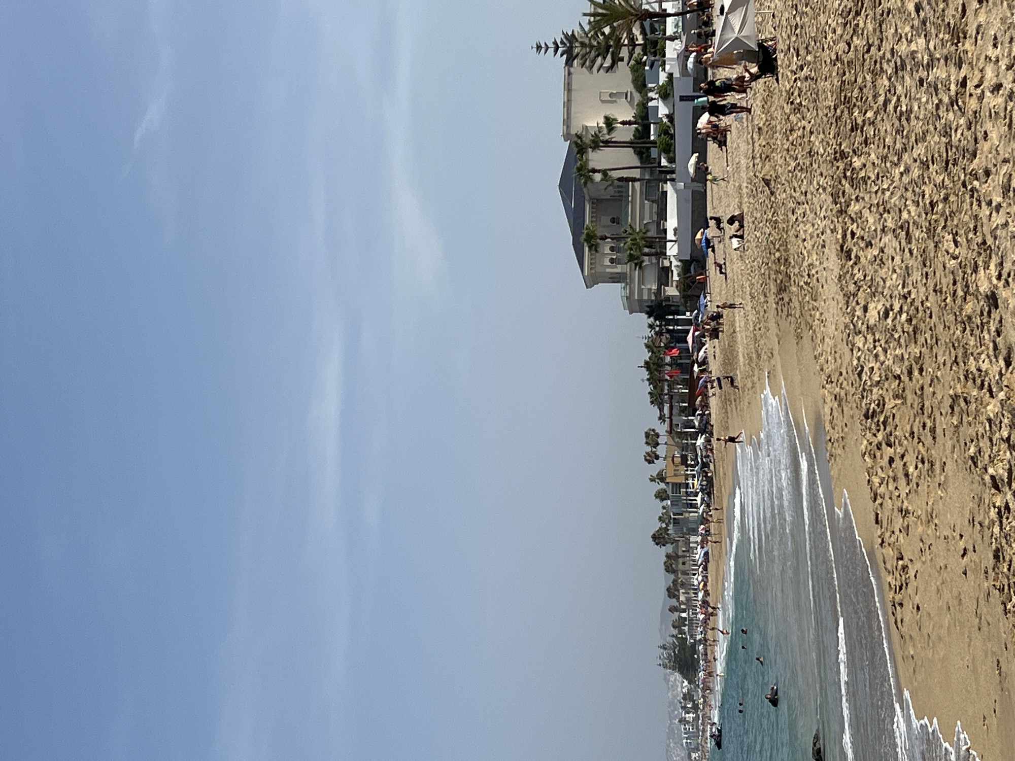 The image depicts a crowded beach in M'Diq, Morocco, on a clear, bright day, likely midday given the strong sunlight casting shadows. The scene is a vibrant panorama of leisure activity, with a long stretch of sandy beach bordering a calm, turquoise sea.
Numerous people are engaged in various beach activities. Some are sunbathing on the sand, dotting the beach with colorful umbrellas providing shade. Others are strolling along the shoreline, while several individuals are in the water, swimming and enjoying the gentle waves. A few are even seen riding a jet ski, indicating a mix of relaxation and water sports.
Behind the beach, a line of modern buildings, including hotels and residences, forms a backdrop. These structures are interspersed with lush palm trees, adding to the tropical ambiance. The architecture suggests a developed tourist area. The sky is a clear, pale blue with a few wispy clouds, contributing to the overall bright and sunny atmosphere.
The overall impression is one of a lively summer day at a popular coastal resort, filled with people enjoying the sun, sand, and sea. The image captures a sense of vacation and recreation. No visible text is present in the image.