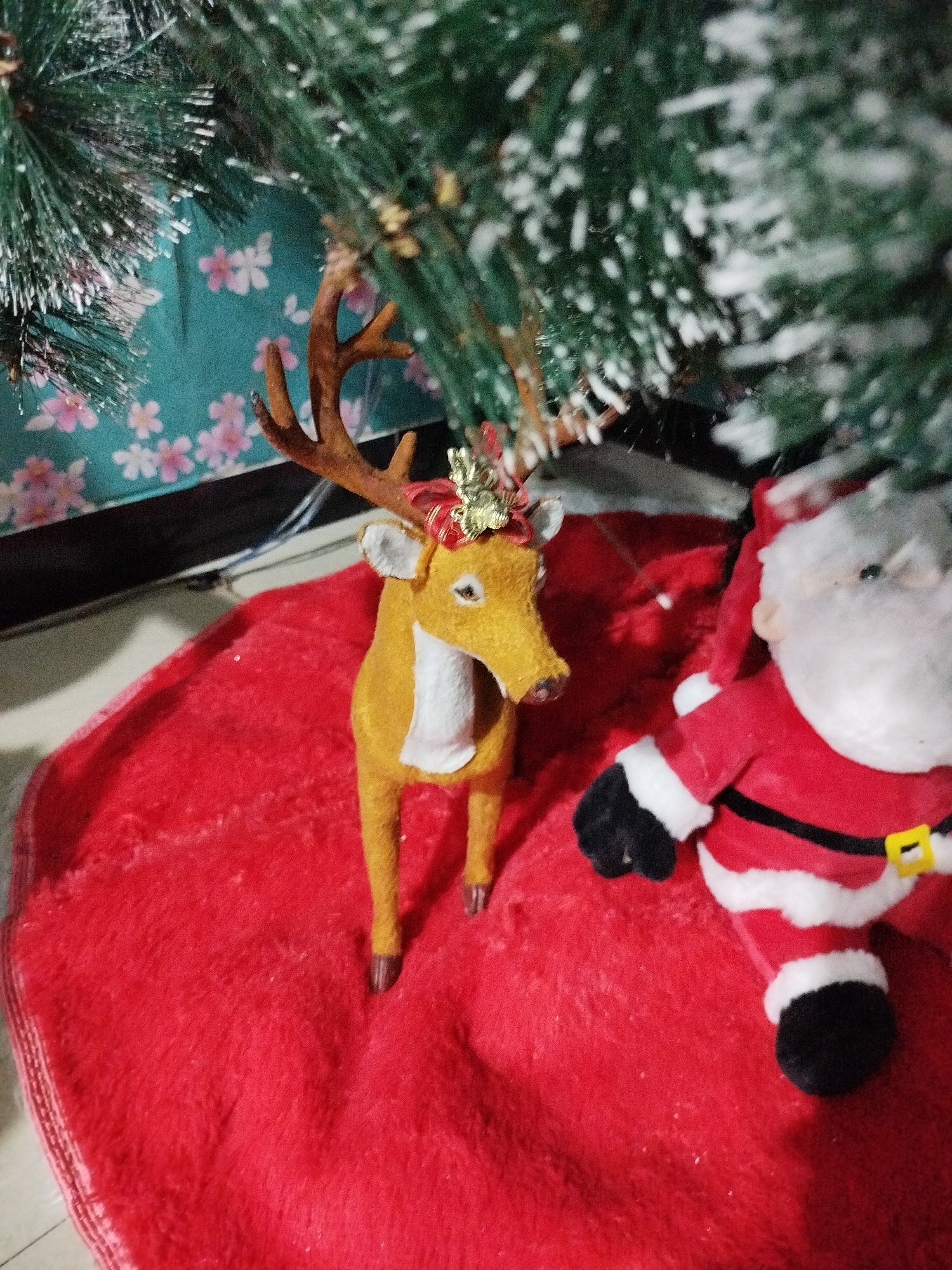 This image depicts a close-up of Christmas decorations. In the foreground, a plush toy reindeer with brown antlers and a red and gold bow sits on a red, fuzzy rug. To the right of the reindeer is a plush toy Santa Claus wearing his traditional red suit with white trim and a black belt. Both toys are positioned under a portion of an artificial Christmas tree, indicated by its green branches dusted with white flocking. The background shows a teal fabric with a pattern of small pink flowers, possibly a tablecloth or curtain. The scene is set indoors, with the floor beneath the rug appearing to be a light-colored tile or linoleum. The overall impression is one of festive holiday decor.