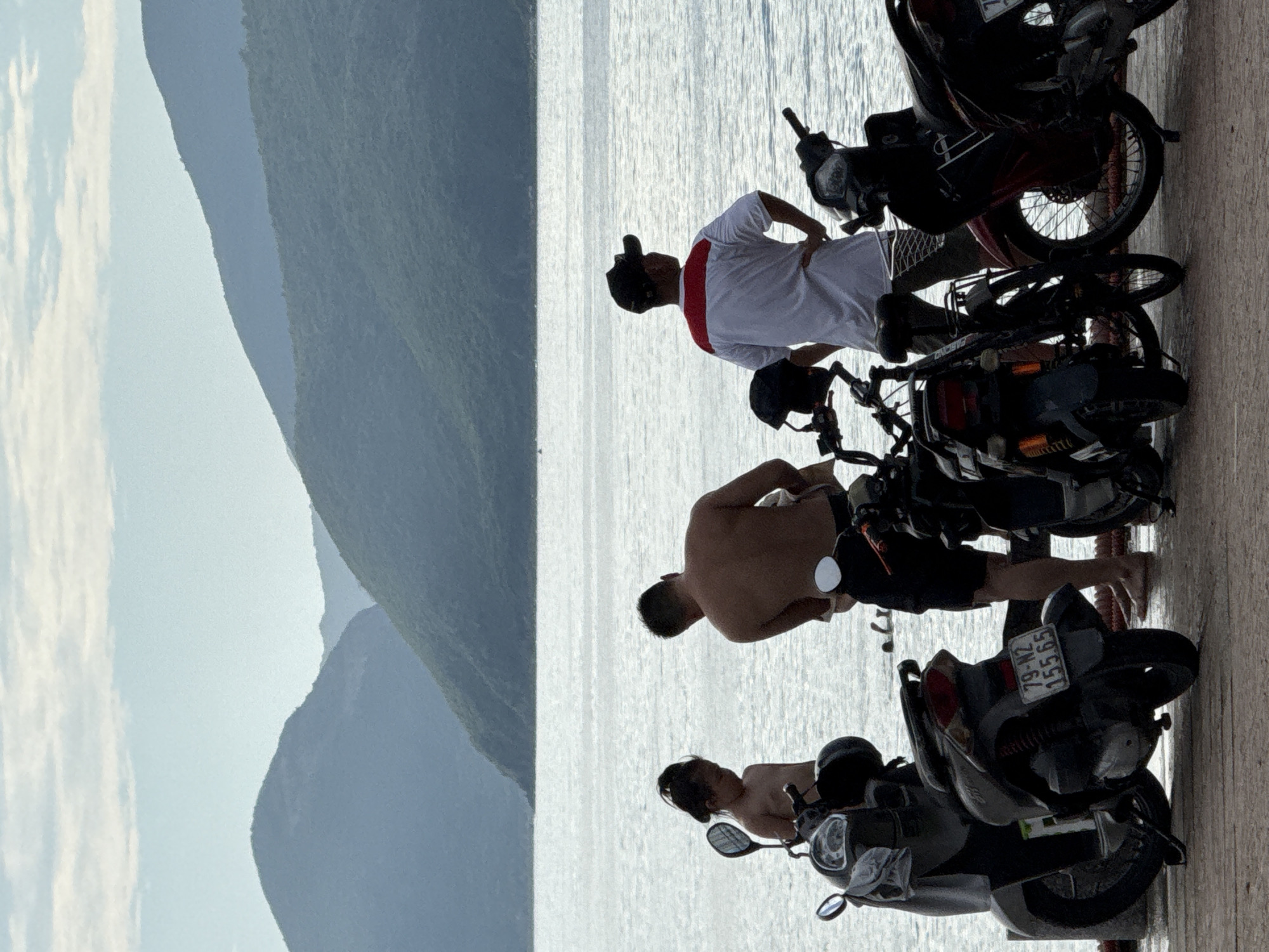 Three individuals, two adult males and one young child, stand near several parked motorcycles on a waterfront promenade. The backdrop features the calm sea and distant, lush green mountains under a cloudy sky. The two adult males are shirtless, one facing away from the camera, looking out at the sea with hands on his hips, wearing a white t-shirt with a red stripe across the chest and a black baseball cap. The other adult male is partially obscured, facing away and looking down. The child, appearing to be a girl, stands beside a motorcycle, looking down. Several motorcycles are parked on the paved surface in the foreground. The sea is calm and reflects the light. The mountains in the distance are layered and covered in dense vegetation. The overall scene suggests a casual stop at a scenic coastal location, likely a common spot for locals and visitors to enjoy the view and the water.
