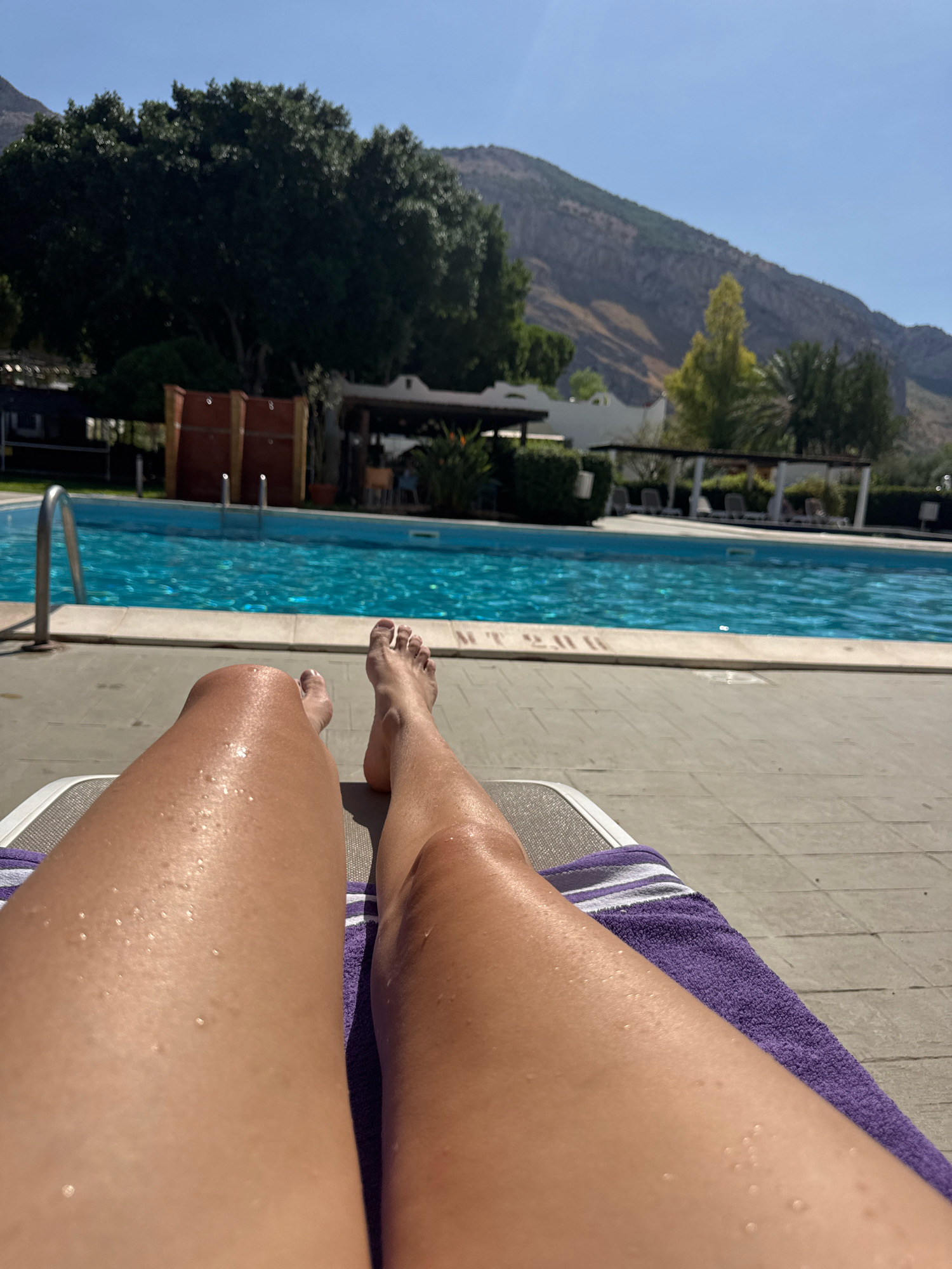A first-person perspective shot shows a person lying on a lounge chair next to a swimming pool. The person's wet legs are visible in the foreground, with water droplets glistening on their skin. A purple towel is draped over the lounge chair. In the background, a bright blue swimming pool is seen, with lounge chairs and umbrellas on the far side. Beyond the pool, there are trees, buildings, and a rocky mountain under a clear blue sky. The overall setting appears to be a resort or hotel in Isola delle Femmine, Italy, given the Mediterranean climate suggested by the presence of the pool and the mountainous landscape.
