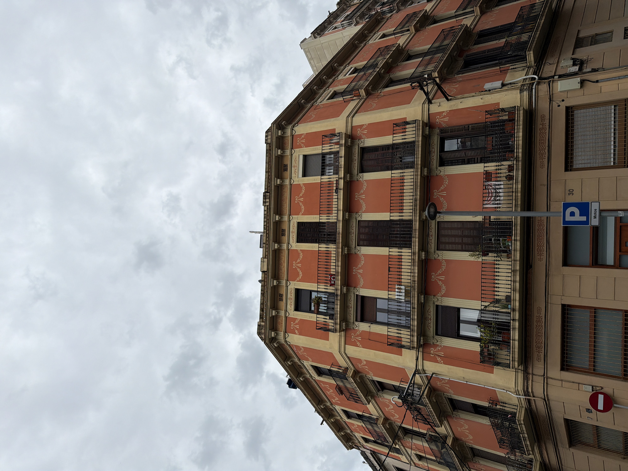 An eye-level shot captures a five-story building in Barcelona, Spain, under a cloudy sky. The building is painted in a terracotta color with cream stripes running horizontally across each floor. The facade is adorned with decorative floral patterns in a lighter shade. Each floor features windows with metal balconies and dark brown shutters, some slightly ajar. A parking sign is visible on the right side of the building. The building is on the narrow street.