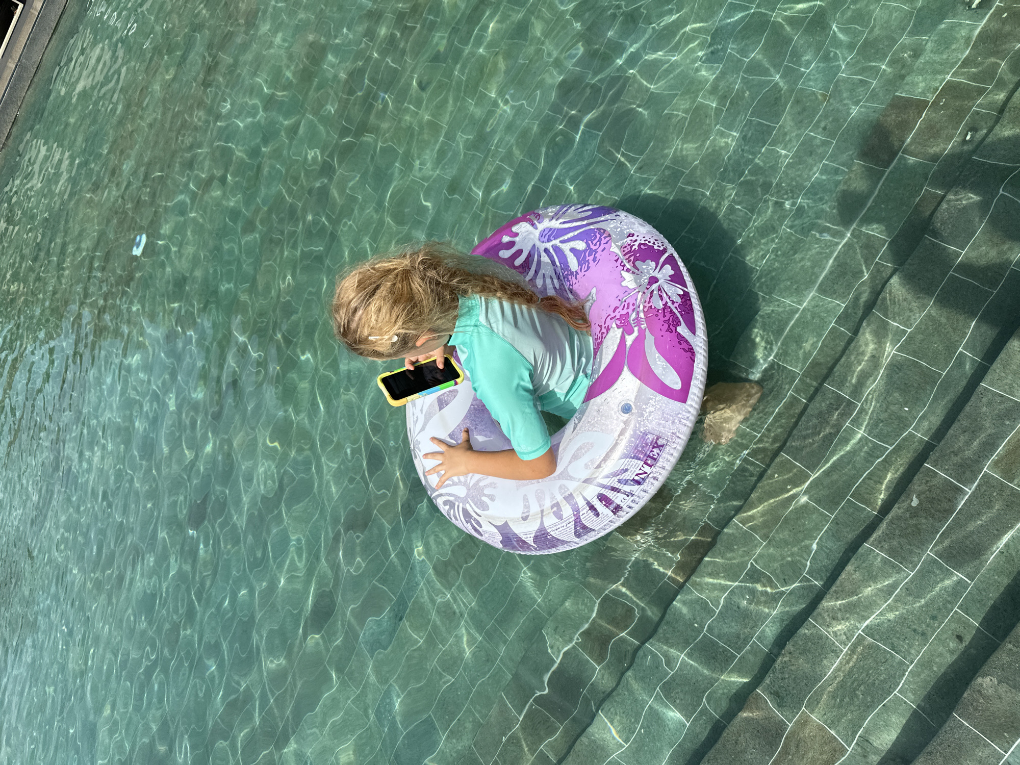 A young Caucasian girl with blonde hair in a braid is floating in a swimming pool on an inflatable ring. She is wearing a turquoise and white rashguard. The girl is holding a yellow-cased phone and appears to be using it. The pool has a tiled bottom and the water is clear and green. Sunlight is creating patterns on the water's surface and the pool's steps. The overall scene is peaceful and suggests a summer day.