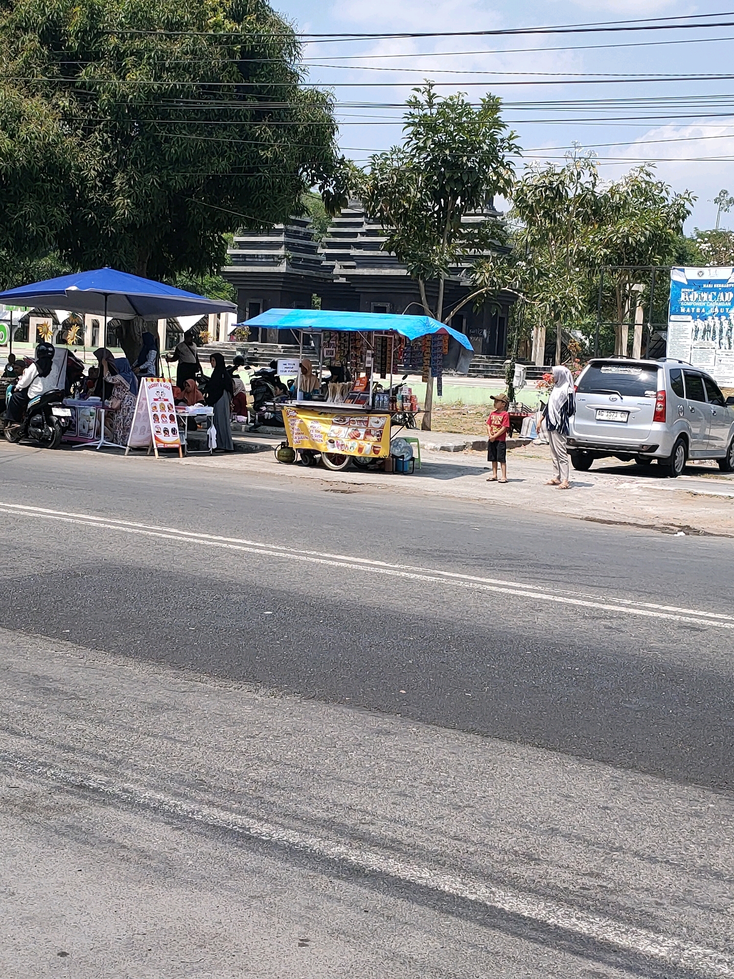 In Kanigoro, Indonesia, a bustling roadside scene unfolds under a bright sky. A silver minivan, license plate AG 1573 GH, is parked on the right side of the road. Two individuals, a woman in a white and blue headscarf and a young boy in a red shirt and black shorts, stand on the sidewalk near the vehicle.
Adjacent to them, a vibrant food cart, adorned with colorful images of food items, is open for business. A young woman in a black headscarf is seen standing next to the cart. To the left, under a large blue umbrella, a small cluster of people are gathered, some seated on motorcycles. A display board with food images is propped up near this group.
Behind the food stalls and umbrellas, a distinctive architectural structure with layered, dark stone roofs is visible, likely a significant building or monument in the area. Lush green trees provide shade, interspersed with power lines stretching across the sky. The road itself is asphalt, marked with white lines, with some visible tire marks suggesting recent activity. A large banner with text, possibly an advertisement or announcement, is affixed to a pole on the far right.