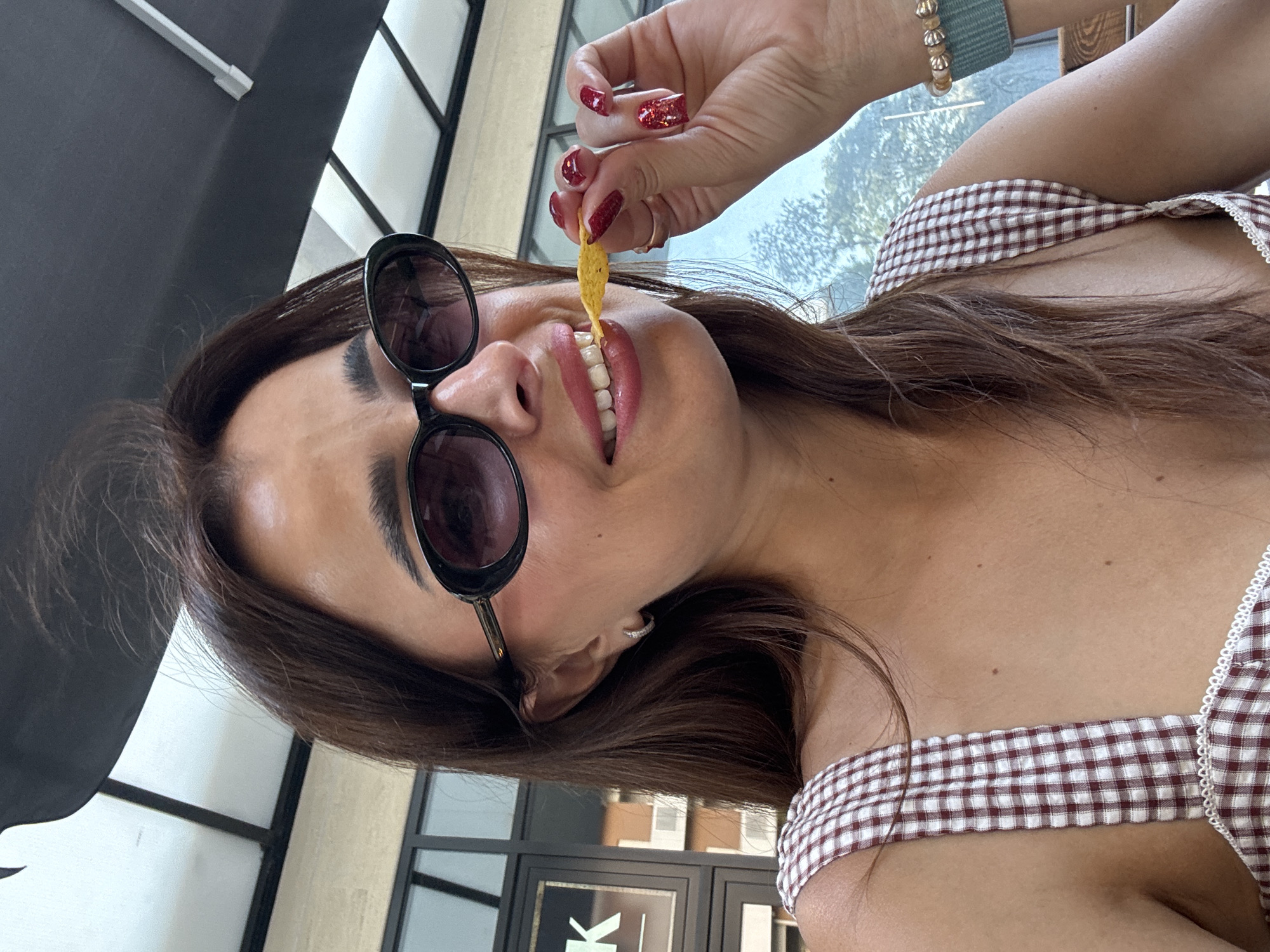 A close-up, eye-level, outdoor selfie captures a woman with medium-length brown hair, wearing black oval sunglasses and a gingham print top, as she eats a chip. The background suggests an urban setting, possibly a cafe or restaurant, with a glimpse of a building and some greenery visible through a window. The overall lighting indicates a daytime scene.