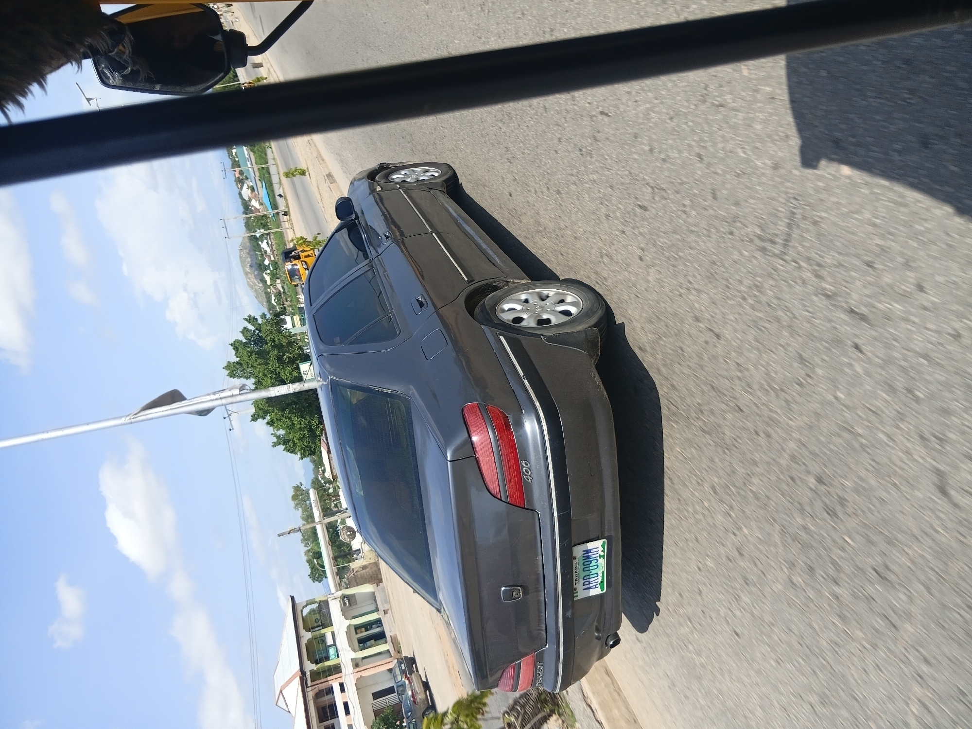 A dark gray Peugeot 406 sedan is parked on a paved road in Jalingo, Nigeria. The car is viewed from the rear-right quarter, with its license plate visible, reading "TARABA ARD-09MM". In the background, a multi-story building with a white and green facade and a light blue roof stands to the left. A tall flagpole with a flag partially unfurled stands in the middle ground. The sky is blue with scattered white clouds. Trees and other buildings are visible further in the background, along with distant hills. The scene appears to be in an urban or semi-urban environment during daylight.