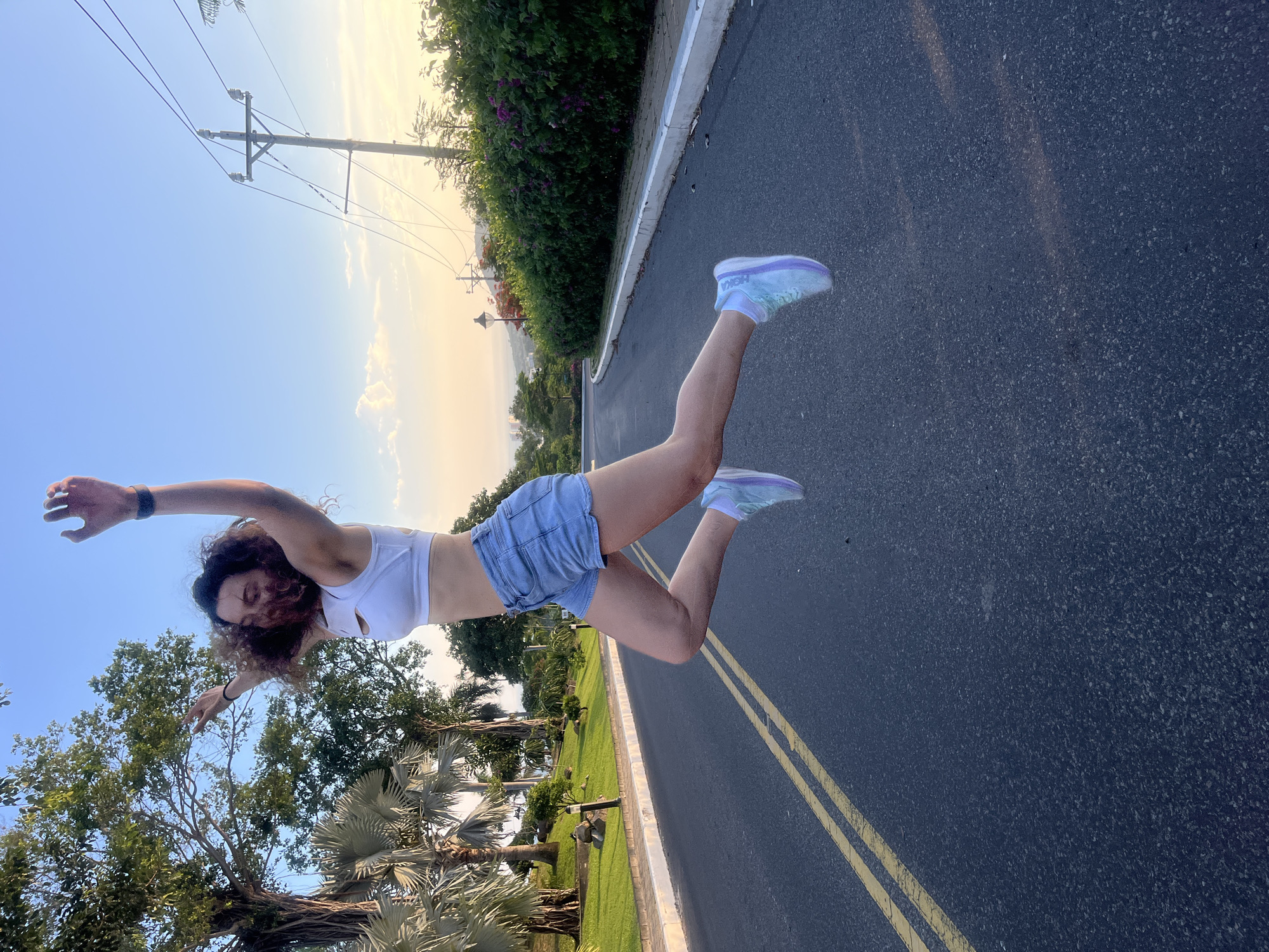 A young woman with curly brown hair is captured mid-air, jumping on a paved road. She is wearing a white sports bra, light blue denim shorts, and multi-colored athletic shoes. Her arms are raised above her head, and her legs are bent as she leaps. The road has a double yellow line in the center and a white curb. To the left of the road, there is a grassy area with palm trees and other foliage, and a streetlamp. To the right, there is a dense hedge with purple flowers, a utility pole with wires, and more trees. The sky is clear and blue, with a hint of sunset colors near the horizon. The location is identified as Phu Thuy Ward, Vietnam.