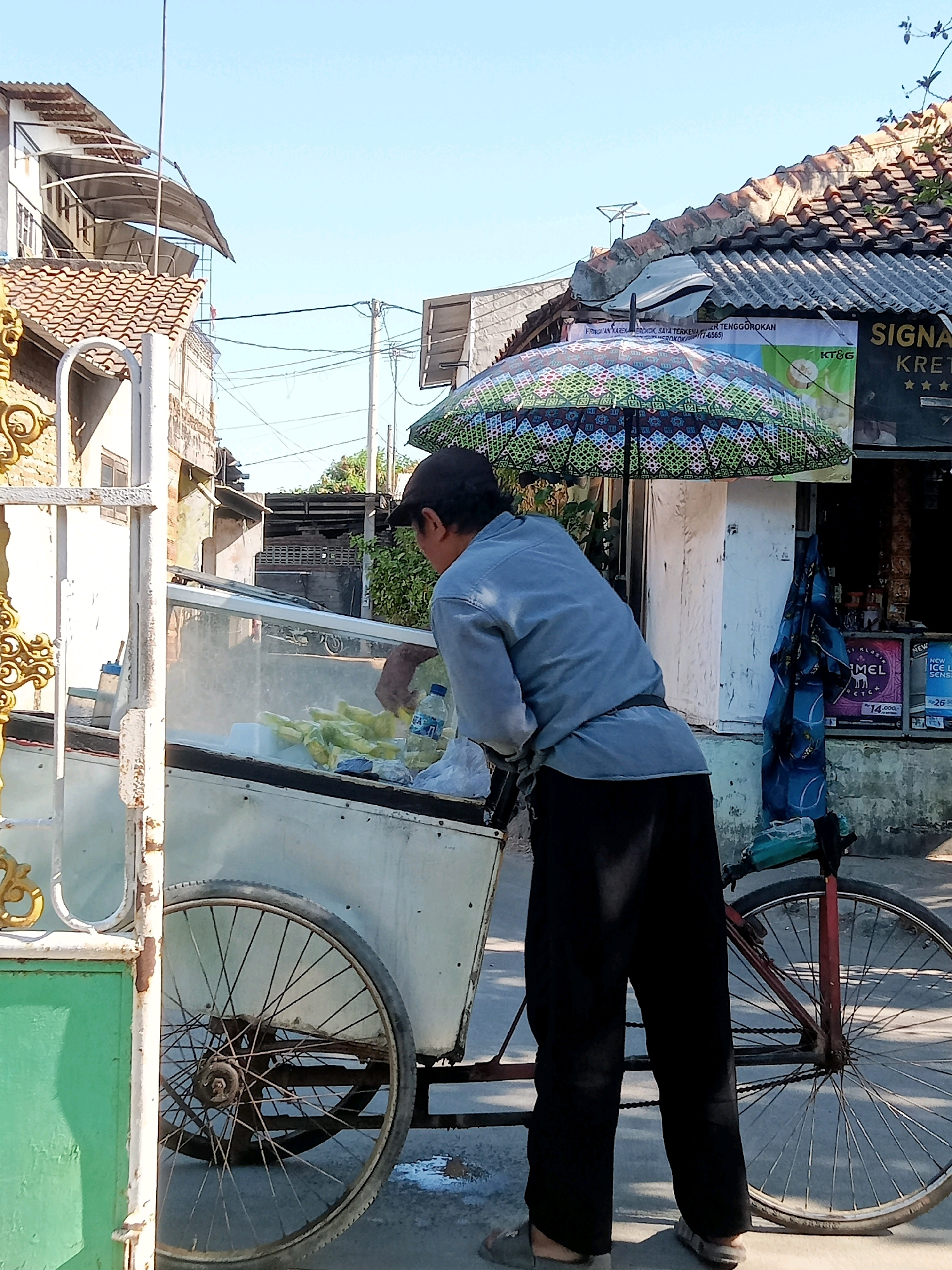 A person wearing a blue shirt, black pants, and a dark baseball cap stands next to a food cart on a street in Cirebon, Indonesia. The cart, which appears to be a pushcart or tricycle cart, has a white body with a glass display case containing green corn. A patterned umbrella with blue, green, and white motifs is open and attached to the cart, providing shade. The person is seen reaching into the cart.
In the background, there are buildings with tiled roofs and some overhead wires. A sign with Indonesian text and logos is visible on one of the buildings. To the right of the cart, a red bicycle wheel is prominent. The ground is paved. The lighting suggests it is daytime, with a clear blue sky. The scene depicts a street vendor preparing or selling food.